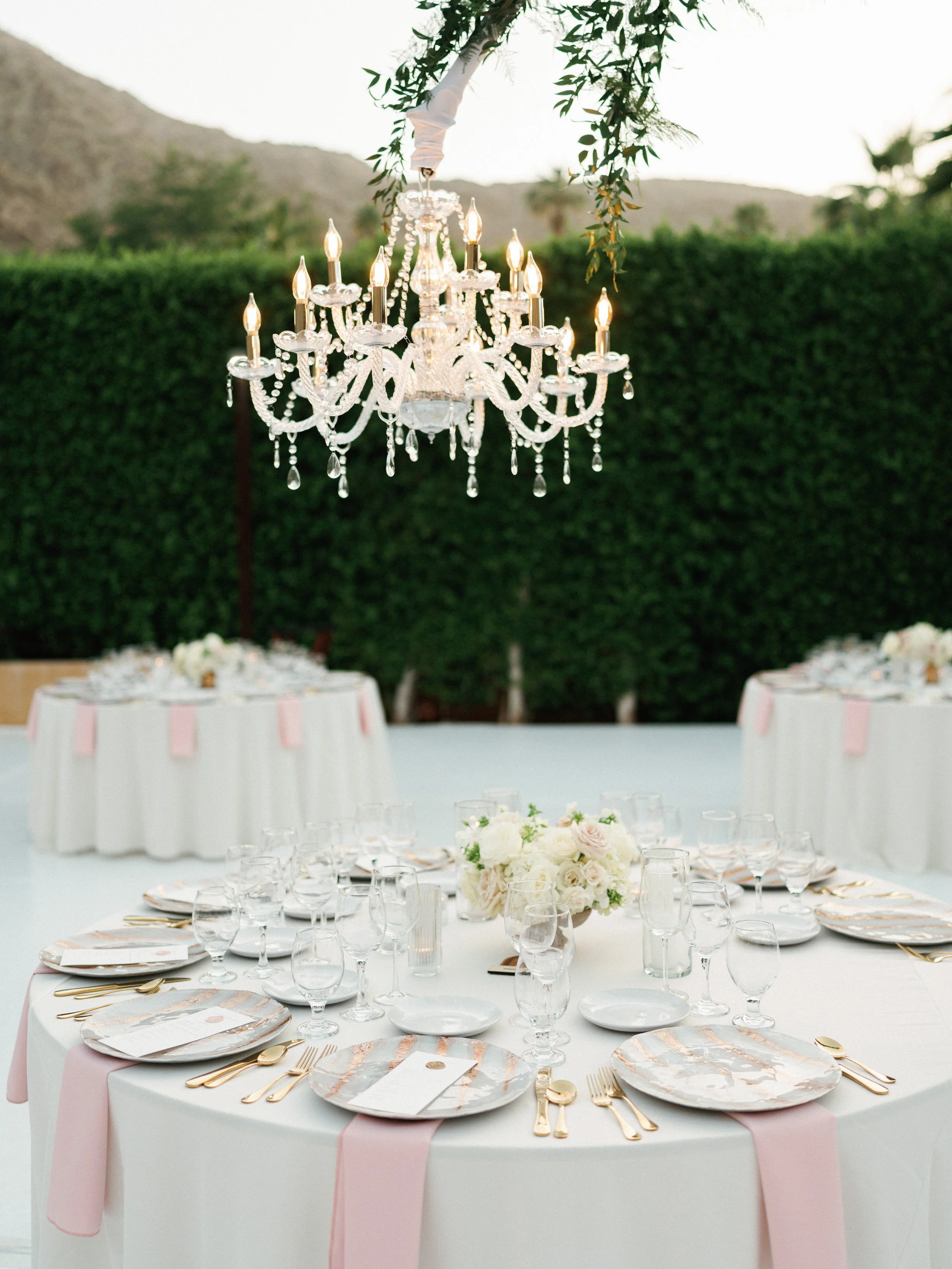 Elegant outdoor wedding reception setup with a long table decorated with white and pink accents, gold flatware, and a floral centerpiece of white roses. A chandelier is hanging above the table, surrounded by greenery.