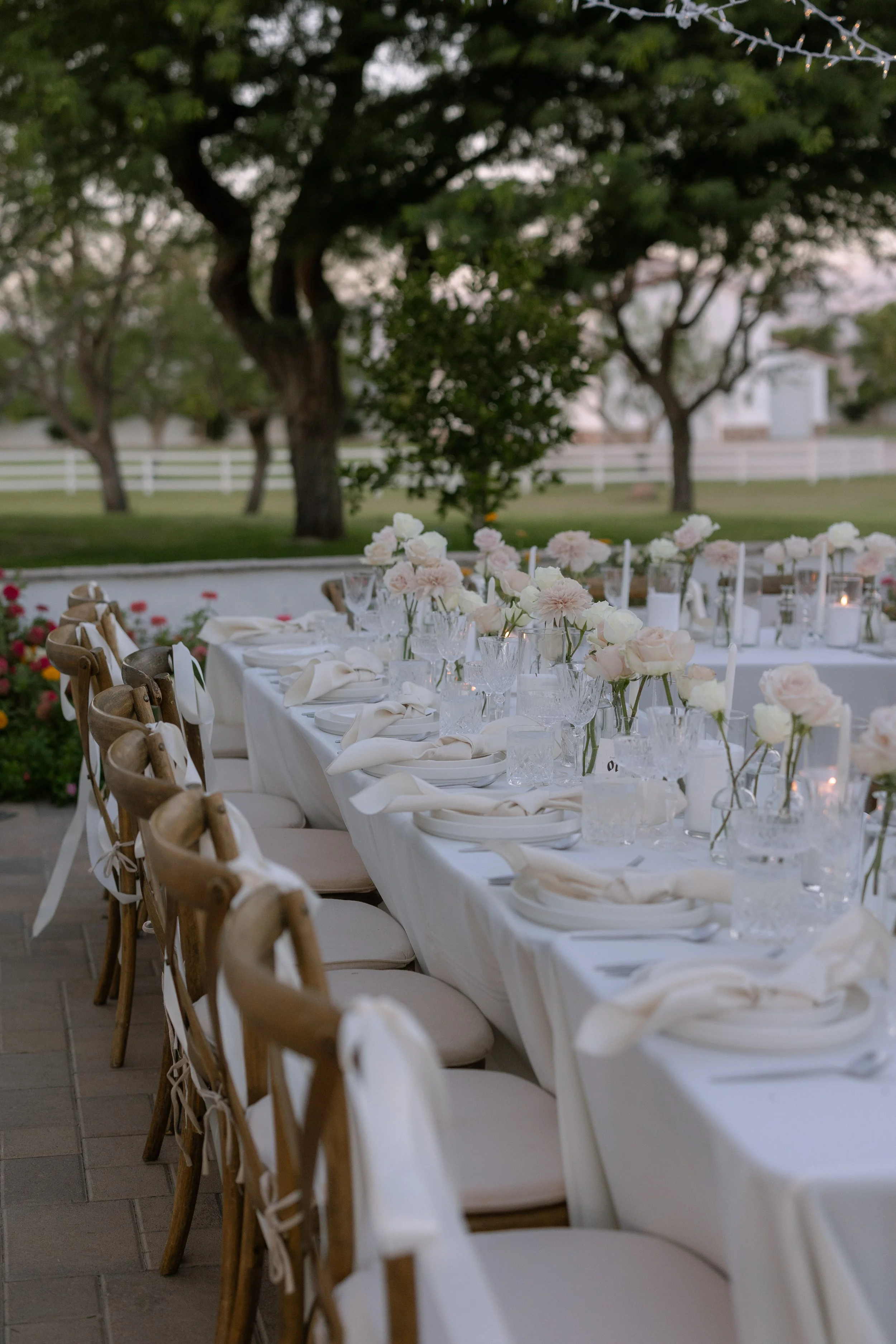 Elegant outdoor dining table setup with white tablecloths, pink and white floral centerpieces, glassware, white napkins, and wooden chairs, set in a garden with trees in the background.