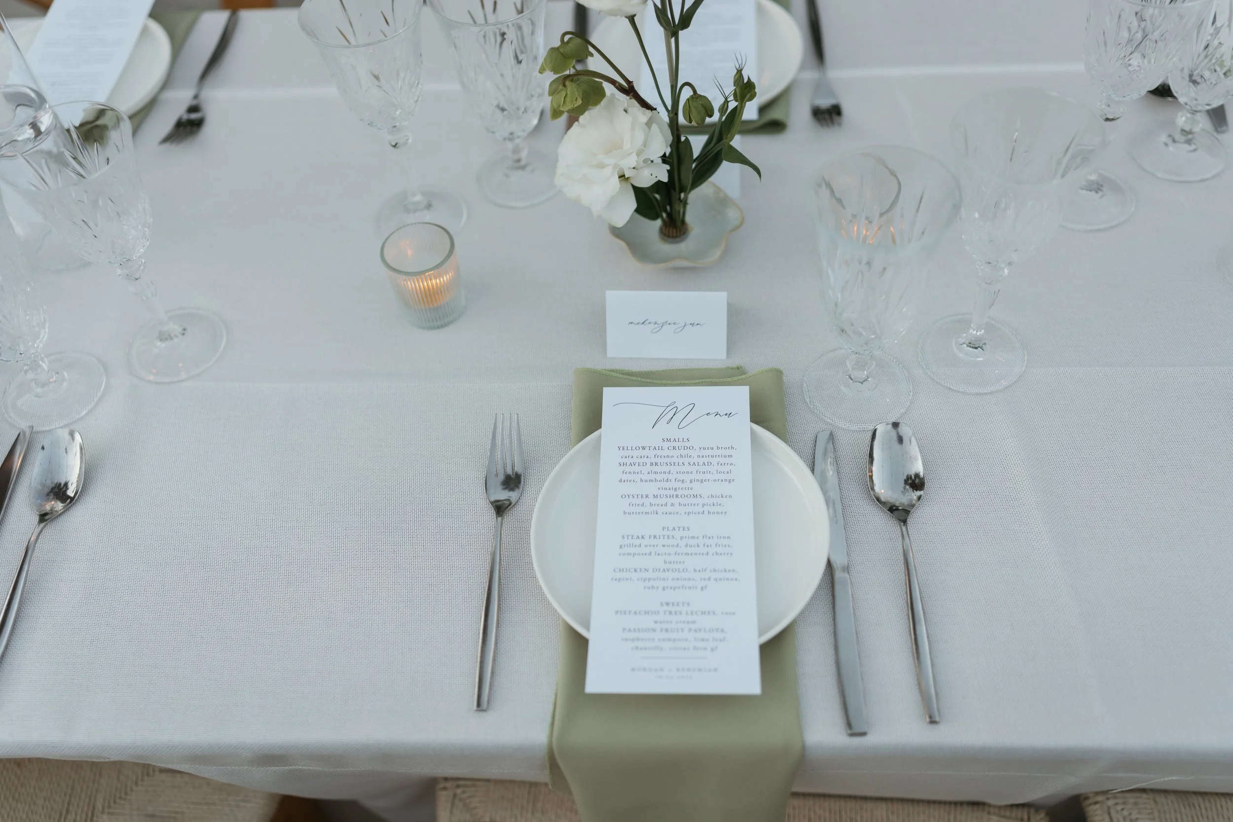 Elegant table setting with multiple wine glasses, cutlery, a floral centerpiece with white flowers, a candle, place cards, and a printed menu on a white tablecloth for a formal event, likely a wedding reception.