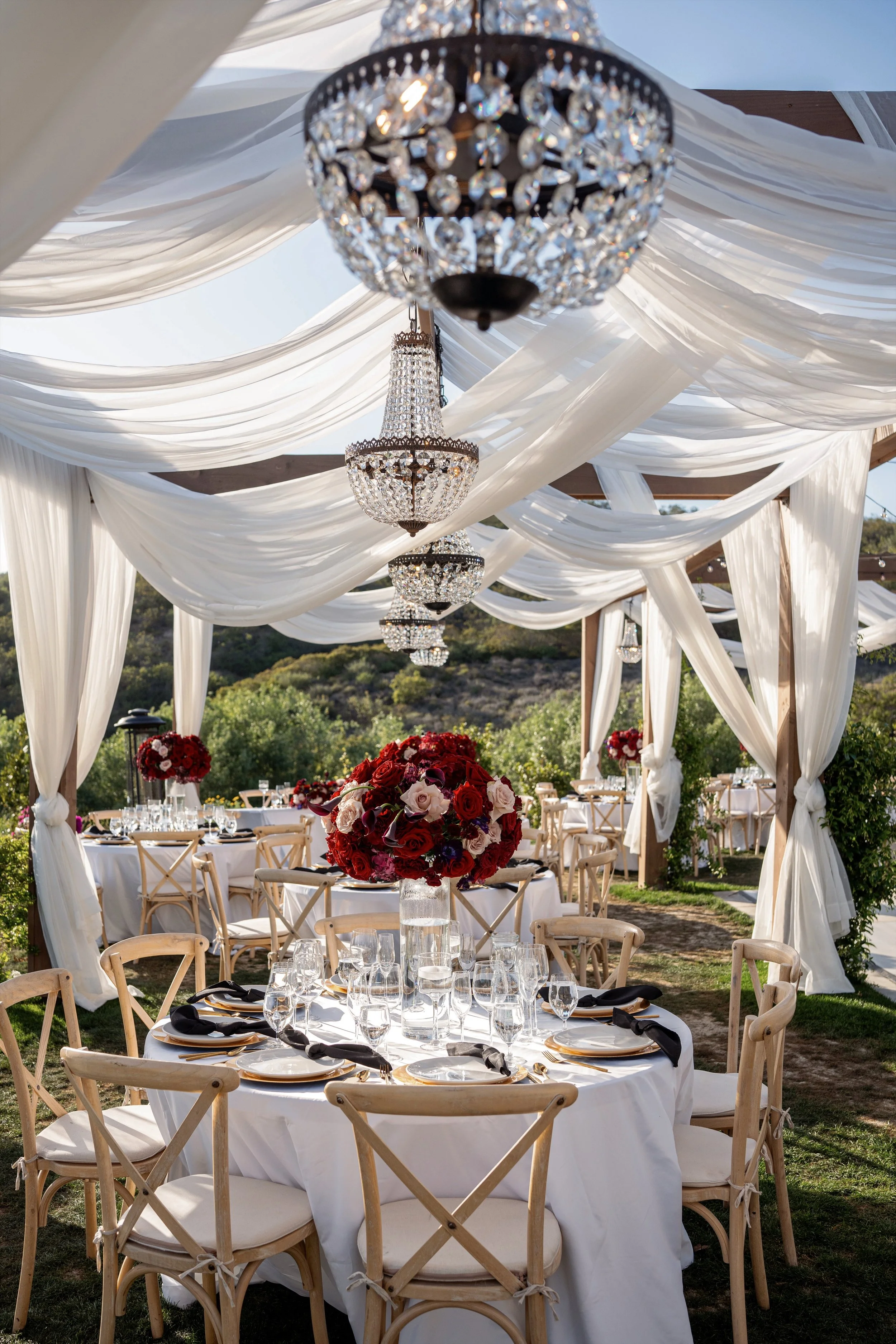 Elegant outdoor wedding setup with round tables covered in white tablecloths, decorated with tall floral centerpieces of red and pink roses, under a canopy with white draped fabric and chandeliers hanging from the ceiling, with a natural landscape in