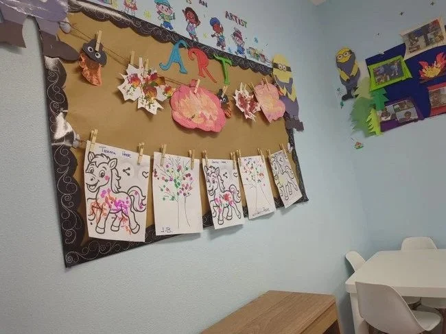 Classroom bulletin board displaying children's artwork, with colorful paper flowers and the word 'ART' in multicolored letters, along with hanging drawings of unicorns and trees.