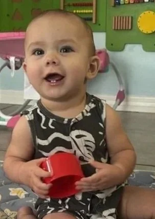 Young child sitting on the floor, smiling, holding a red toy cup in a playroom with colorful furniture and wall decorations.