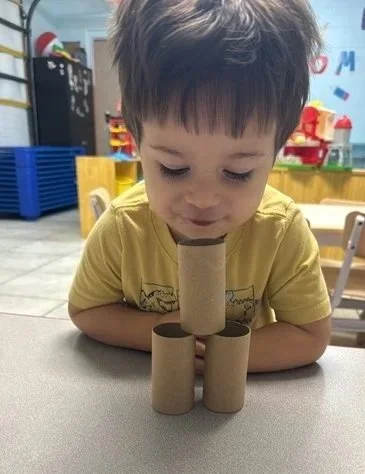Child with dark hair looking down at a tower of four cardboard toilet paper rolls on a table in a colorful classroom.
