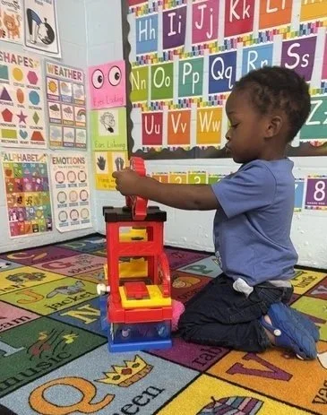 A young boy playing with a stacker toy in a colorful classroom with alphabet posters and educational charts.