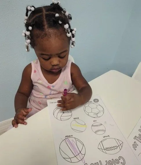 A young girl with braided hair and beads, wearing a pink tank top, coloring a worksheet with geometric shapes at a white table against a light blue wall.