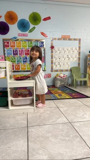A young girl standing in a colorful classroom, smiling and holding a tray in front of a learning wall with alphabet and number charts, decorated with paper fans and a learning clock.