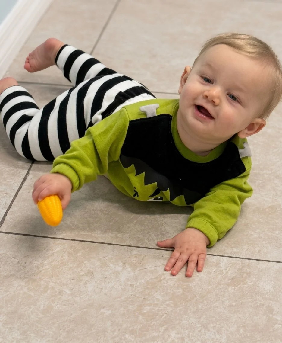 A baby lying on a tiled floor, smiling, wearing black and white striped pants and a green and black Batman shirt, holding a yellow toy in one hand.