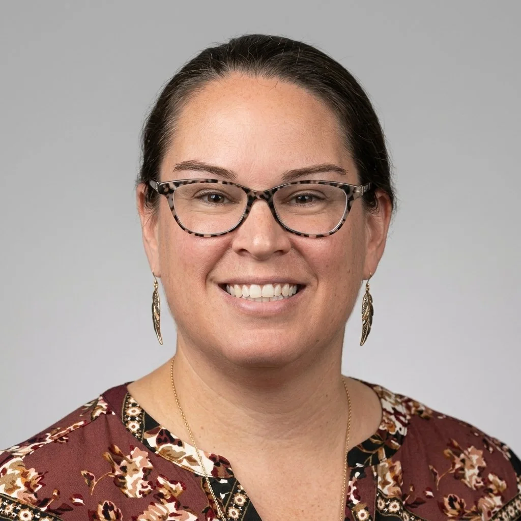 A woman with dark hair, glasses with a tortoiseshell frame, earrings, and a necklace, smiling at the camera. She is wearing a patterned blouse.