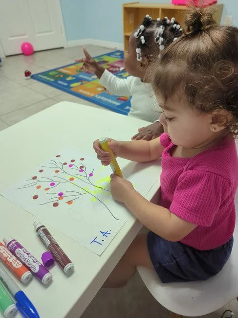 A young girl in a pink shirt drawing a colorful tree on a white paper with markers, with another girl in the background making a peace sign.