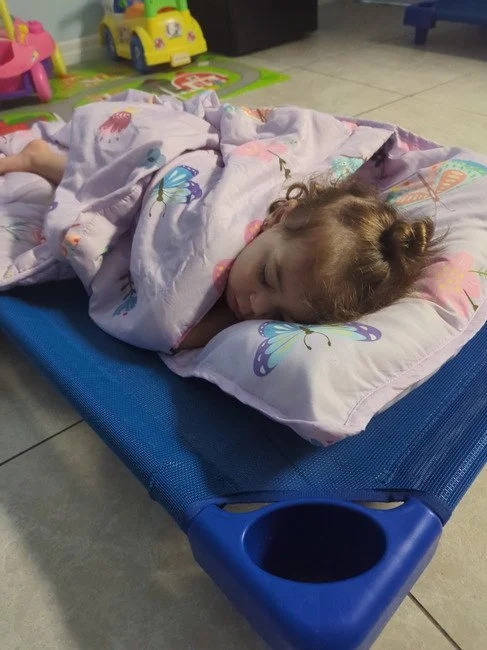 A young girl sleeping on a blue sleep cot with butterfly print pillow, surrounded by children's toys in a playroom.