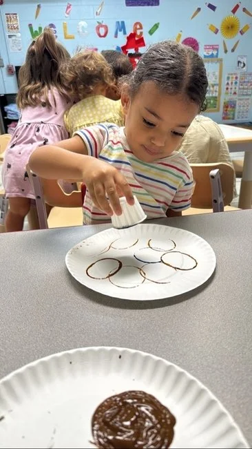 Young girl at a table in a classroom decorates cookies with chocolate sauce, with other children in the background.