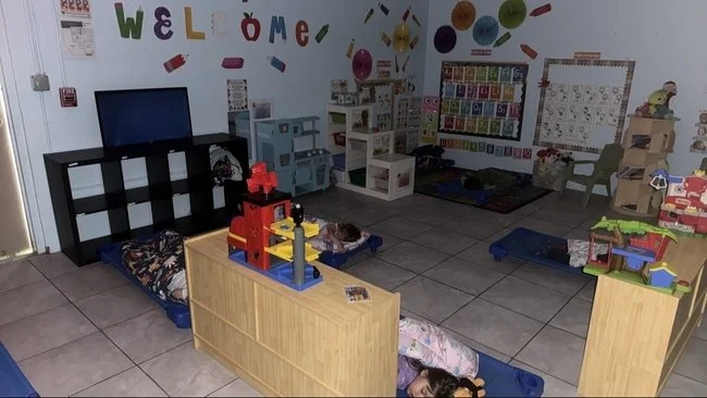 Children napping on mats in a classroom decorated with colorful wall art, alphabet charts, and educational toys.