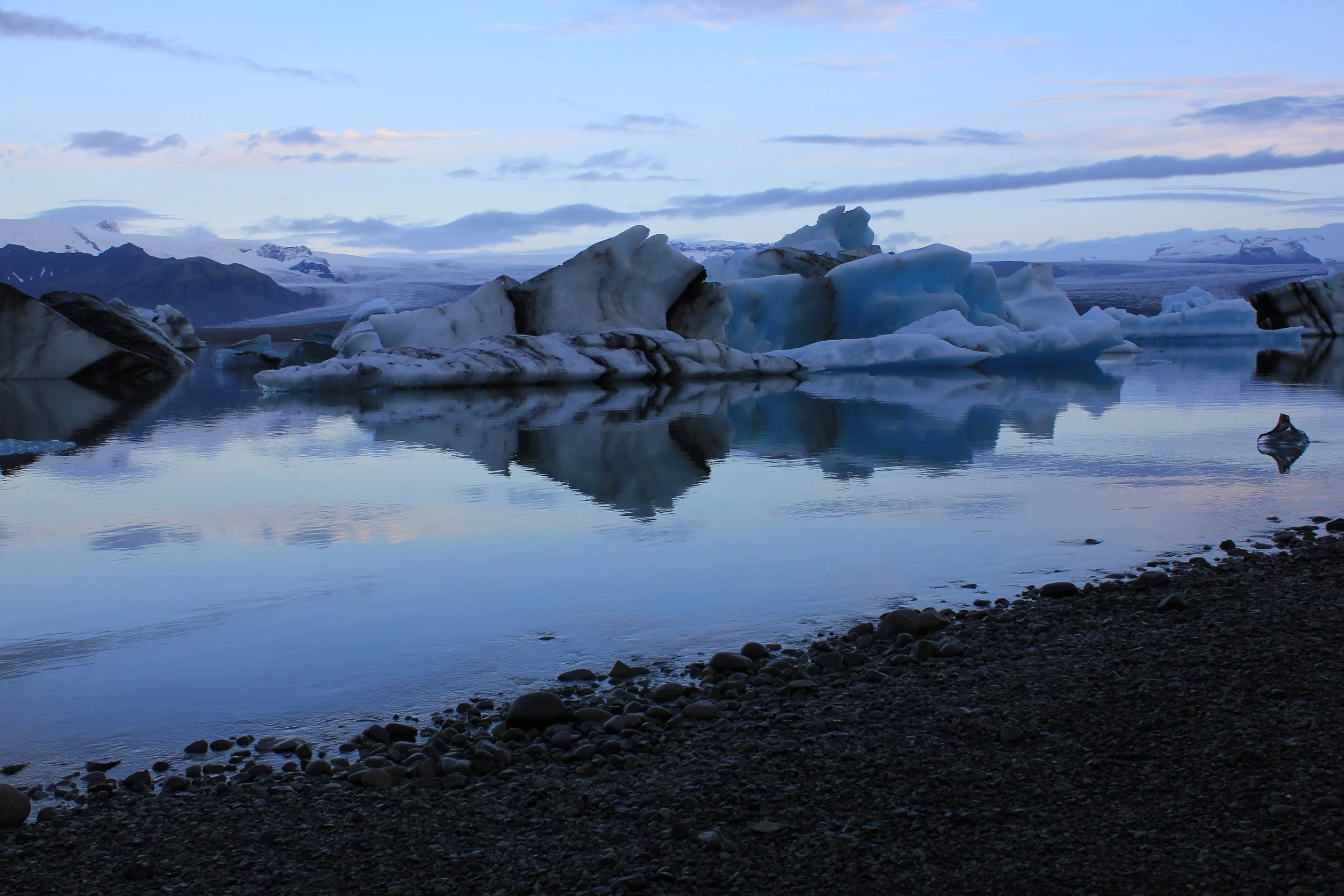 Icebergs floating in a calm, reflective body of water with snow-covered mountains and a cloudy sky in the background.
