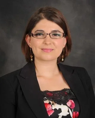 A woman with short brown hair, wearing glasses, a black blazer, and floral top, smiling against a dark gray background.