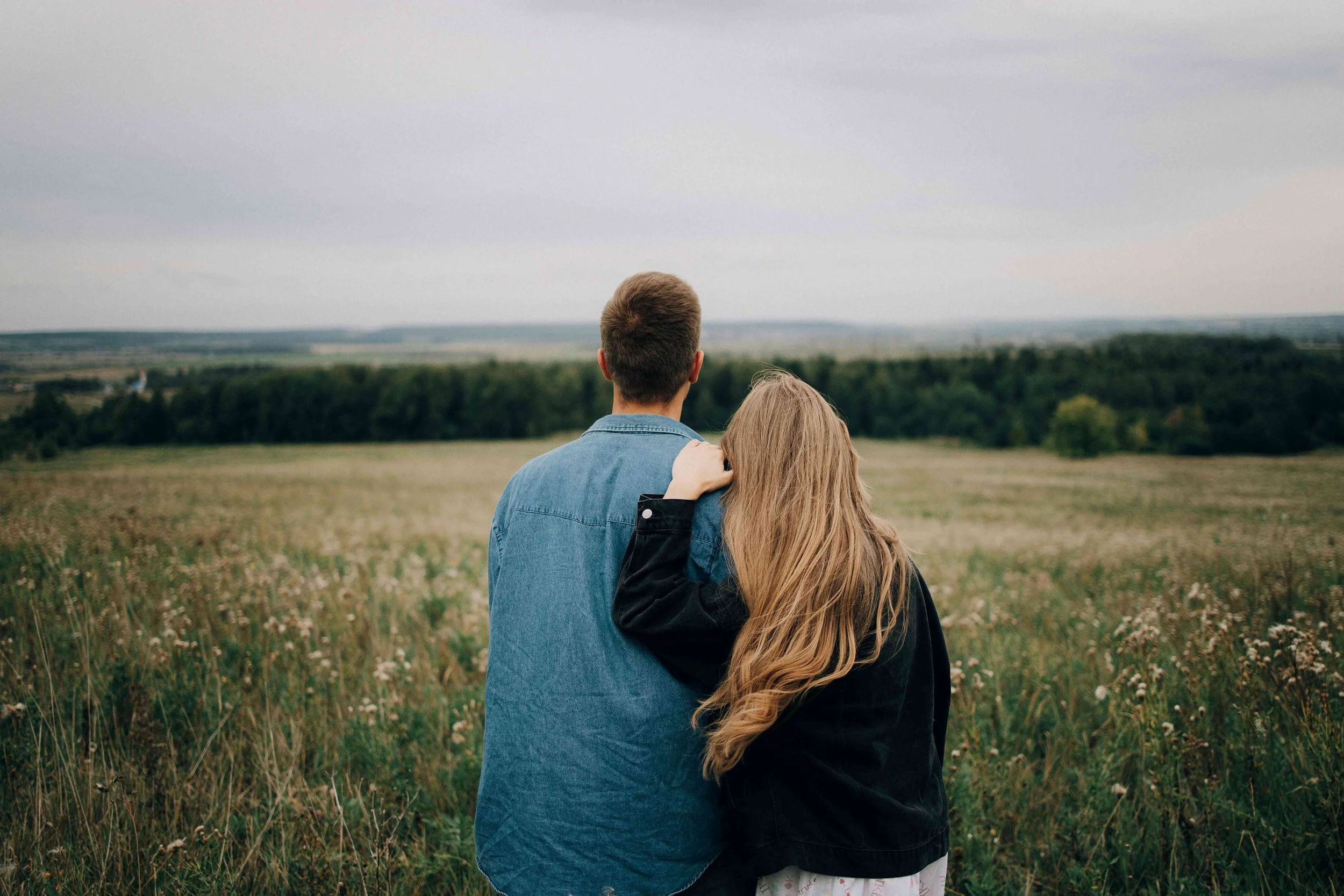 A couple seen from behind standing together in an open field, the woman resting her head on the man's shoulder, gazing toward a wide horizon — representing connection and healing through couples therapy in Los Angeles, CA.