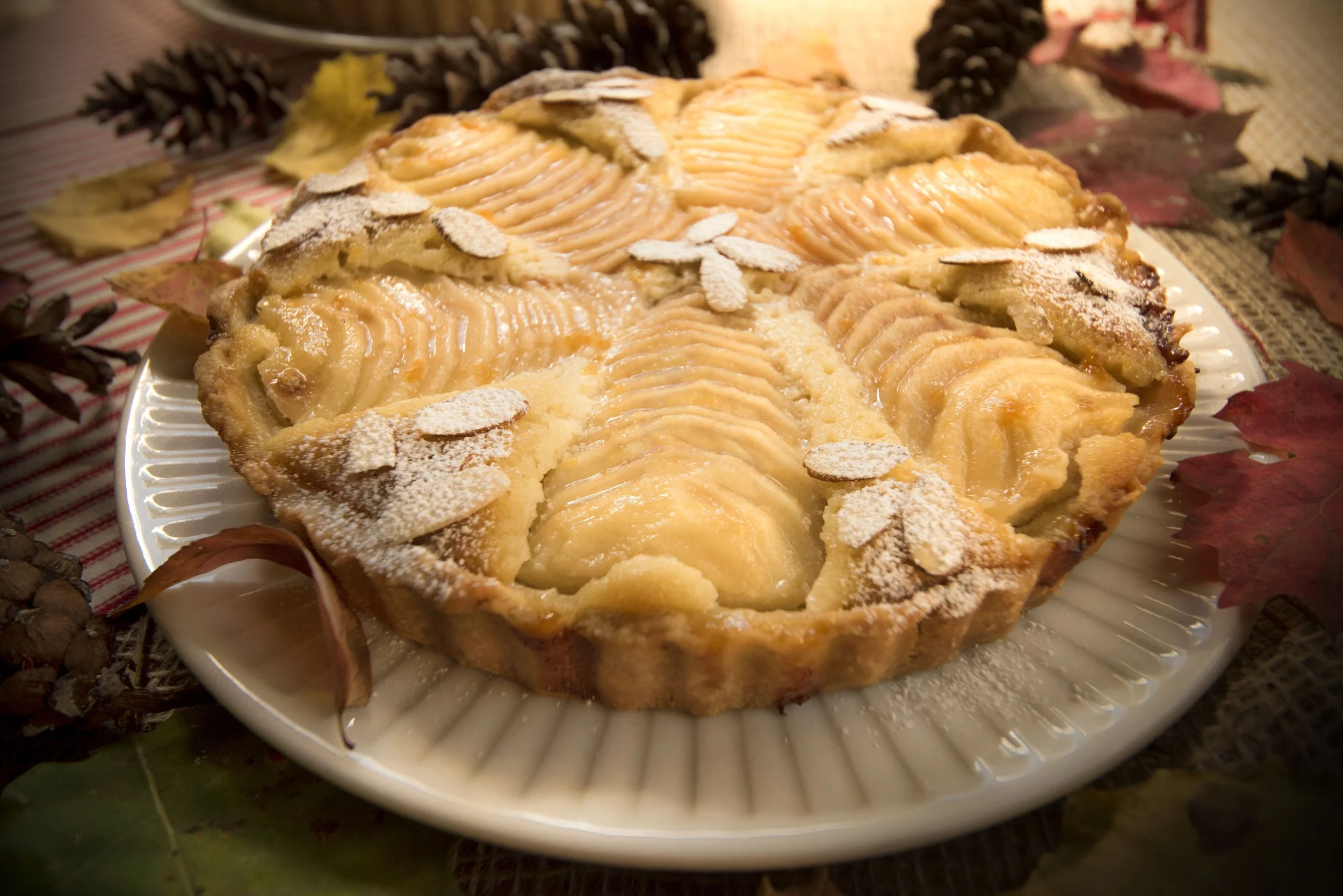 A tart with sliced apples and almond flakes on top, dusted with powdered sugar, placed on a white plate with fall leaves and pinecones around it.
