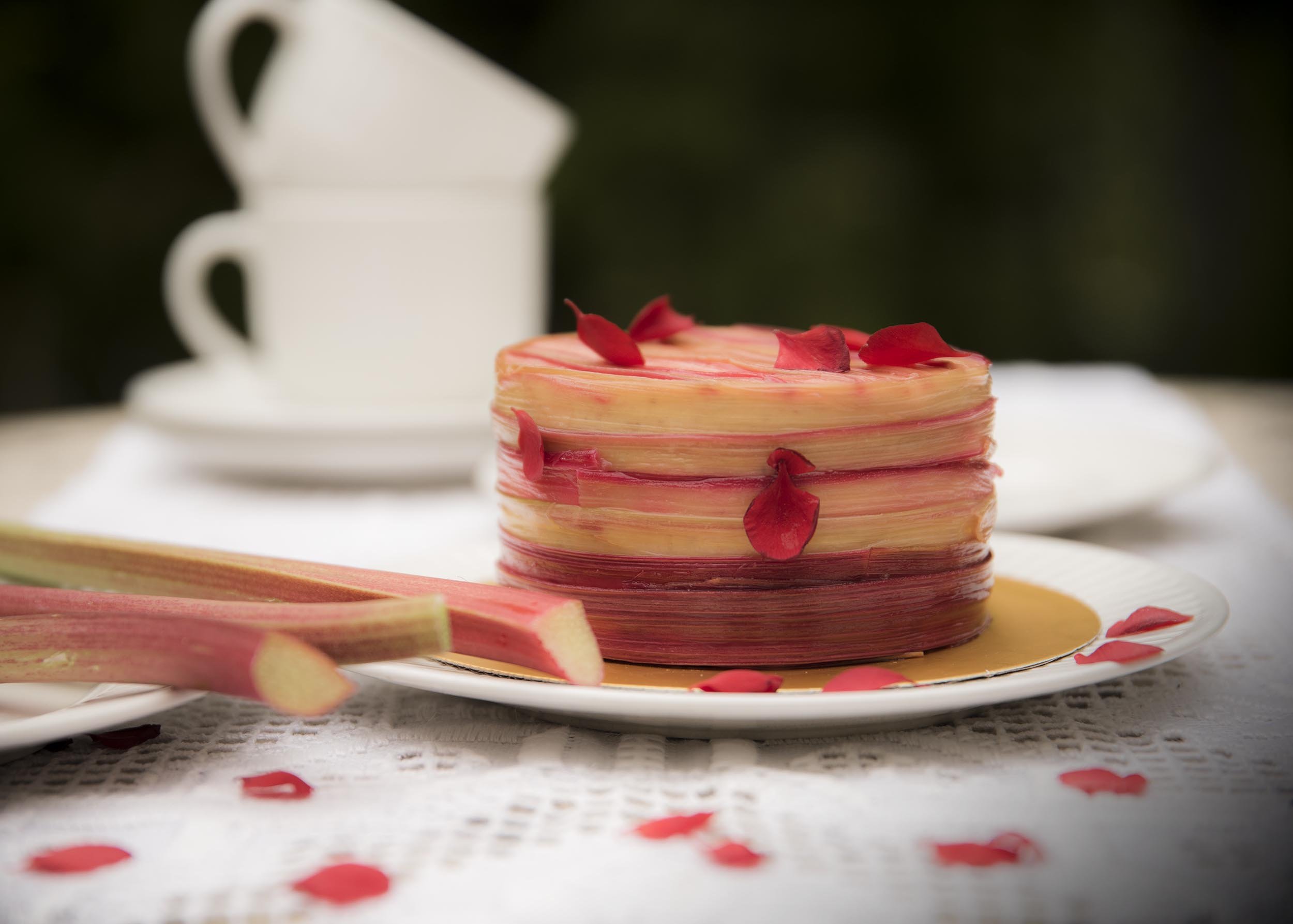 A layered rhubarb cake decorated with red flower petals on top, sitting on a white plate with additional petals scattered around, with a 2 blurred white mugs in the background.