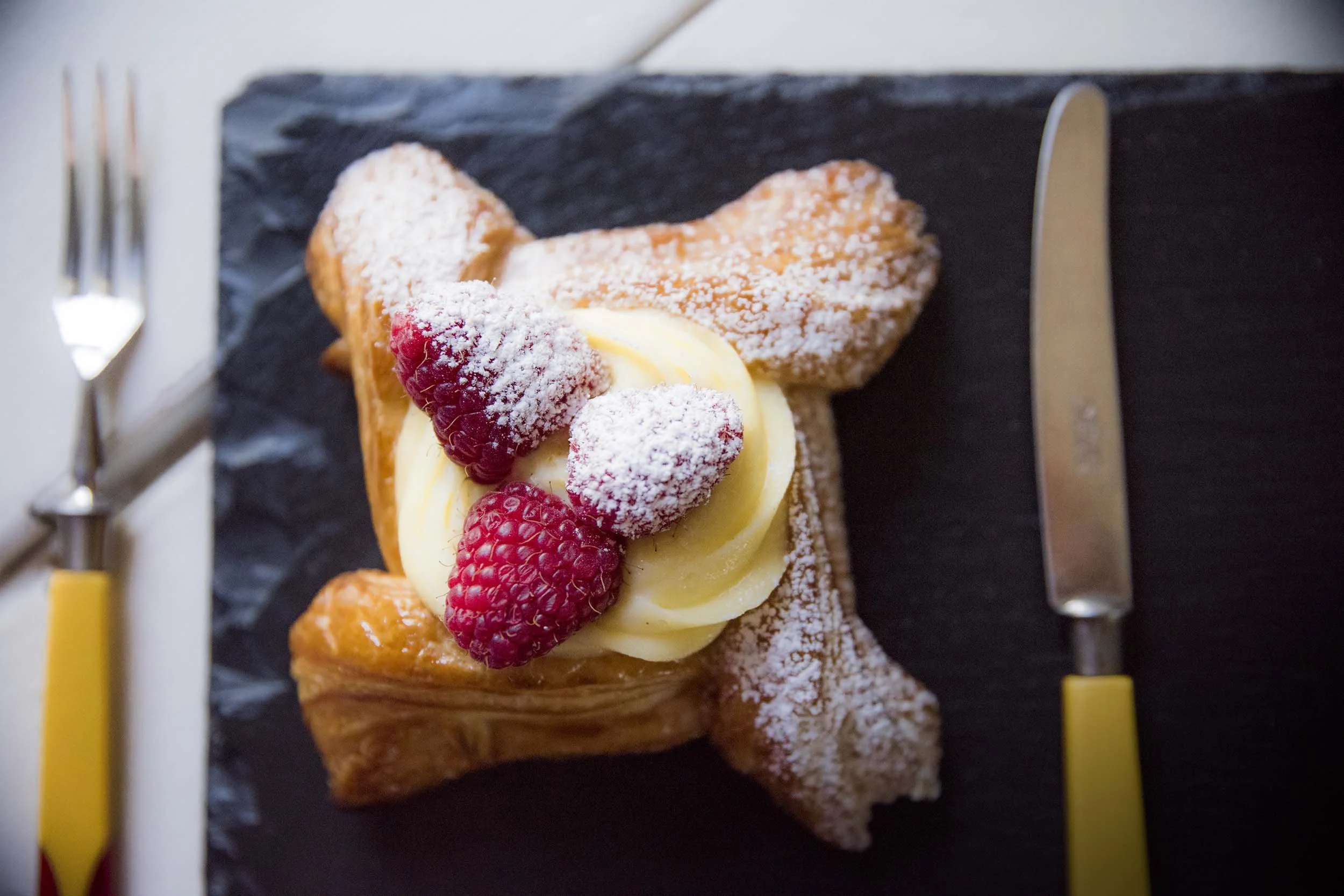 Danish pastry decorated with vanilla custard, raspberries, and powdered sugar, presented on a black slate serving board surrounded by forks and yellow utensils.