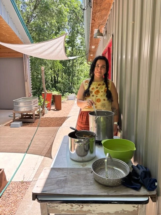 Woman standing next to a table withdye pots and bowls outside a metal building, with trees and a sunny sky in the background.