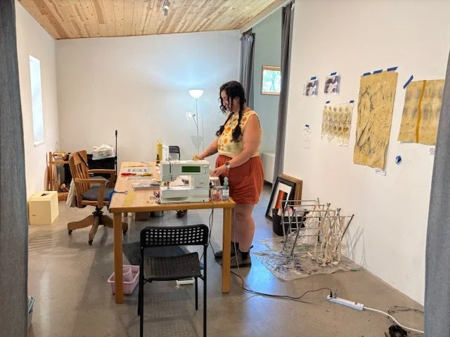 A woman working with fabric and sewing machines in a well-lit room with art on the walls.