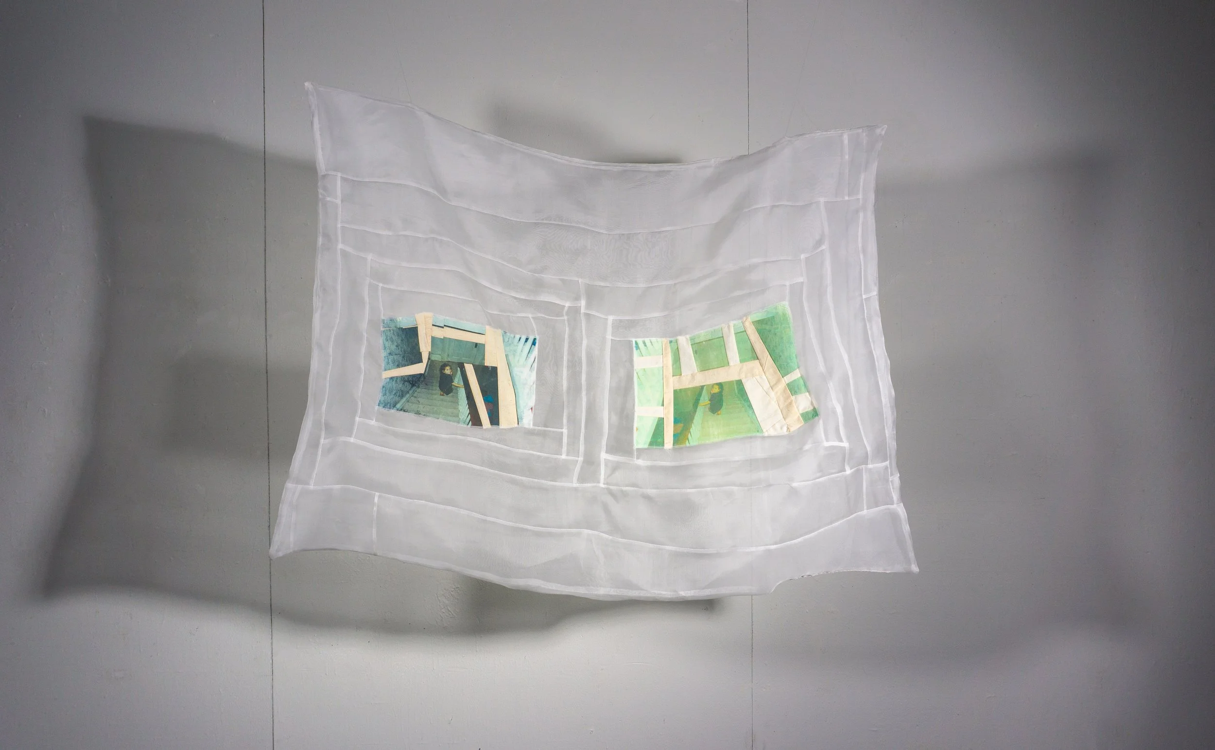 A white, semi-transparent fabric with two printed photos sewn onto it, hanging against a plain wall.
