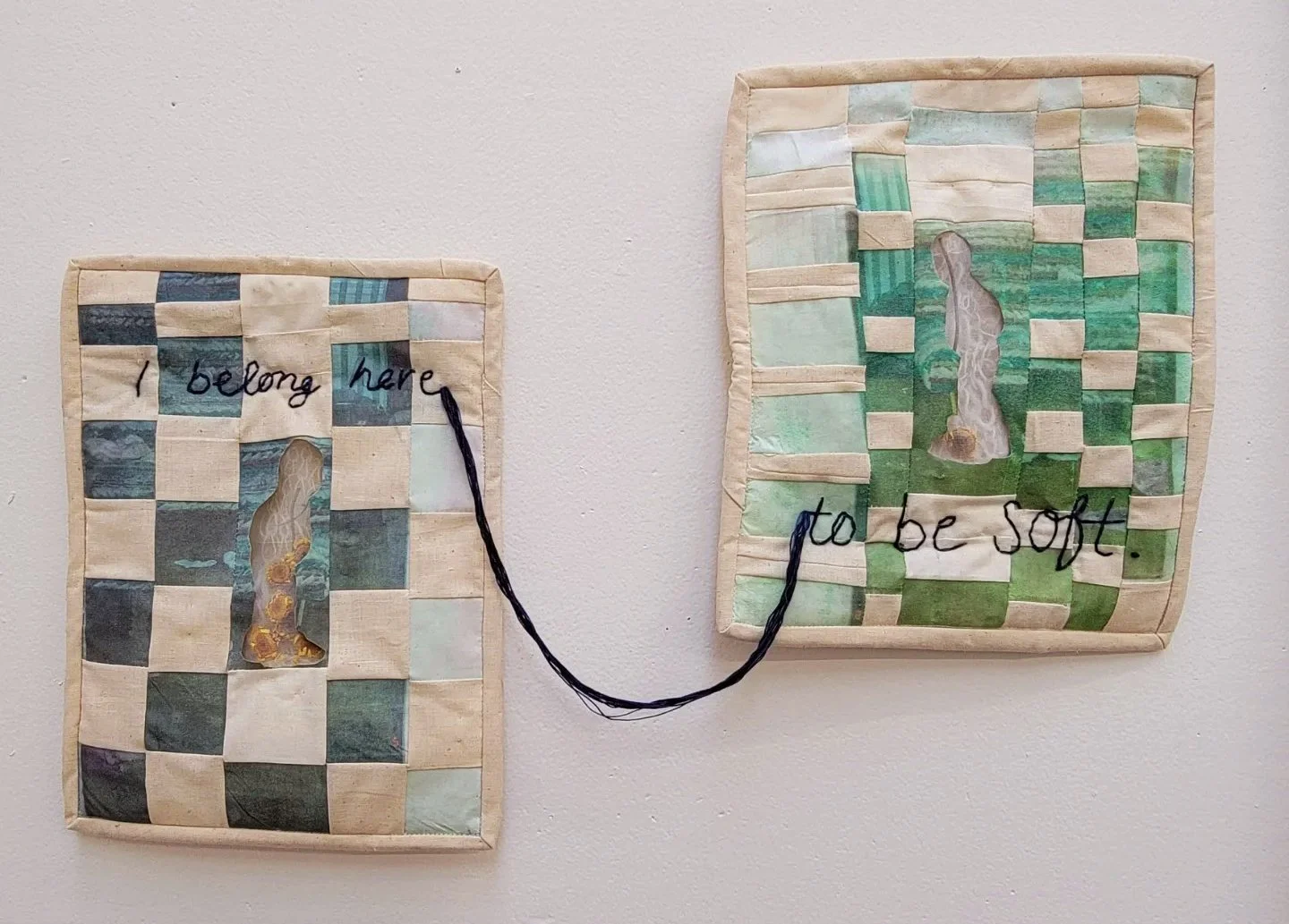 Two quilted fabric squares connected by indigo thread, with hand-stitched embroidered text. The left quilt reads 'I belong here' and the right quilt reads 'to be soft'.