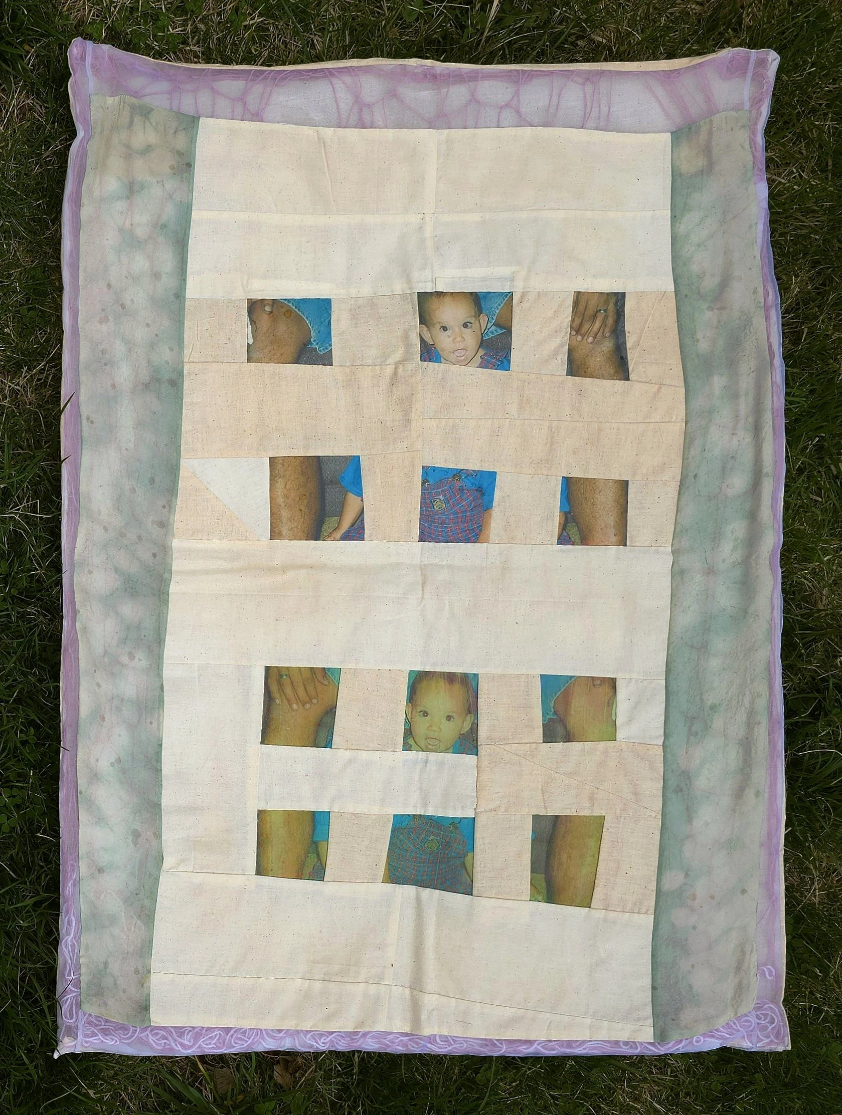 A fabric quilt with a pieced grid of cut-out photo transfers of a child and father's adult hands, laid on grass outside.