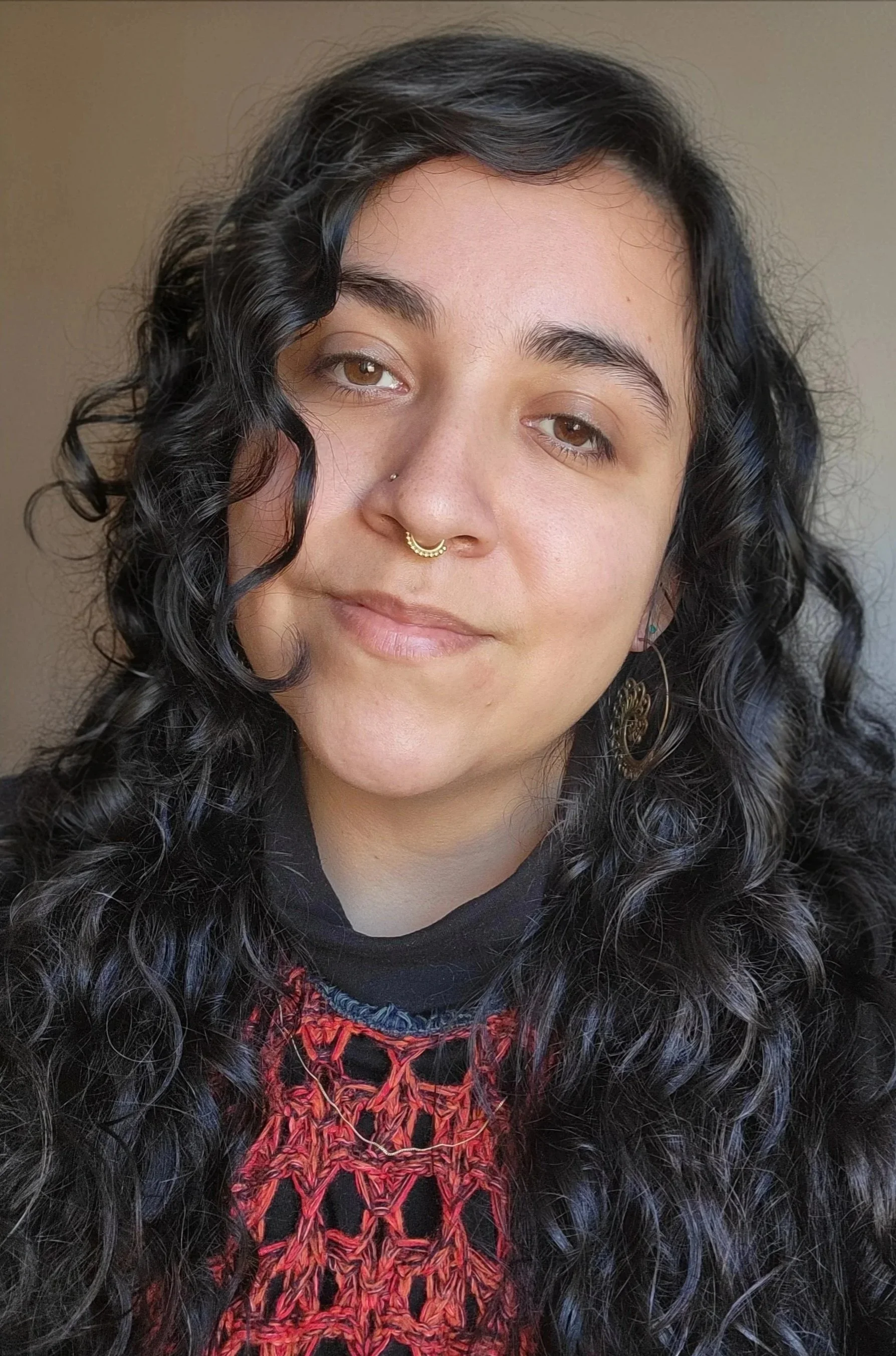 Close-up portrait of a young woman with long, curly dark brown hair, brown eyes, a septum piercing. She is wearing earrings and a black top with a red and black woven pattern.