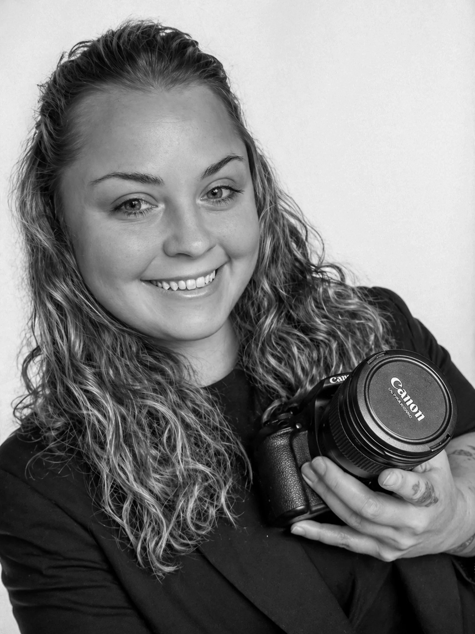 Black and white portrait of smiling woman with curly hair holding a Canon camera.