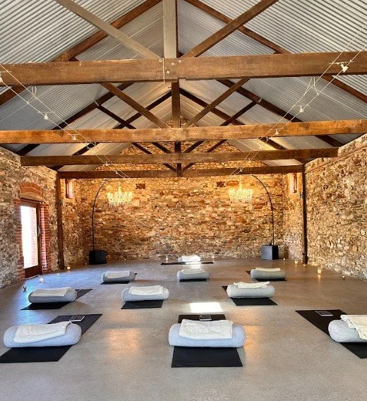 A spacious yoga studio with a high ceiling and exposed brick walls, arranged with nine mats, each with a towel and bolsters, set for a class. Chandeliers hang from the ceiling, and sunlight enters through small windows.