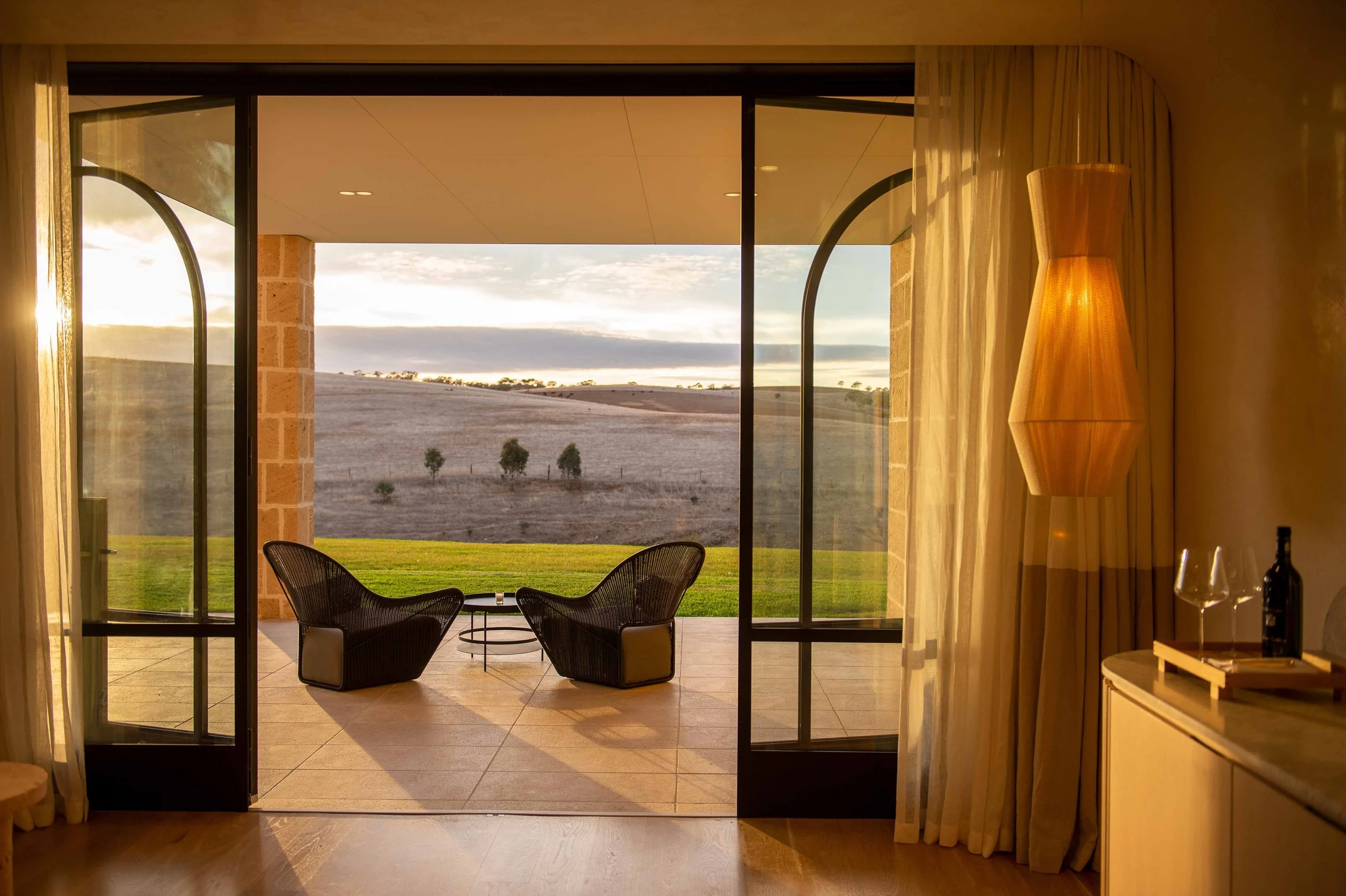 Living room with sliding glass doors opening to a patio and countryside view; two black chairs and a small round table outside, gold floor lamp, and curtains inside.