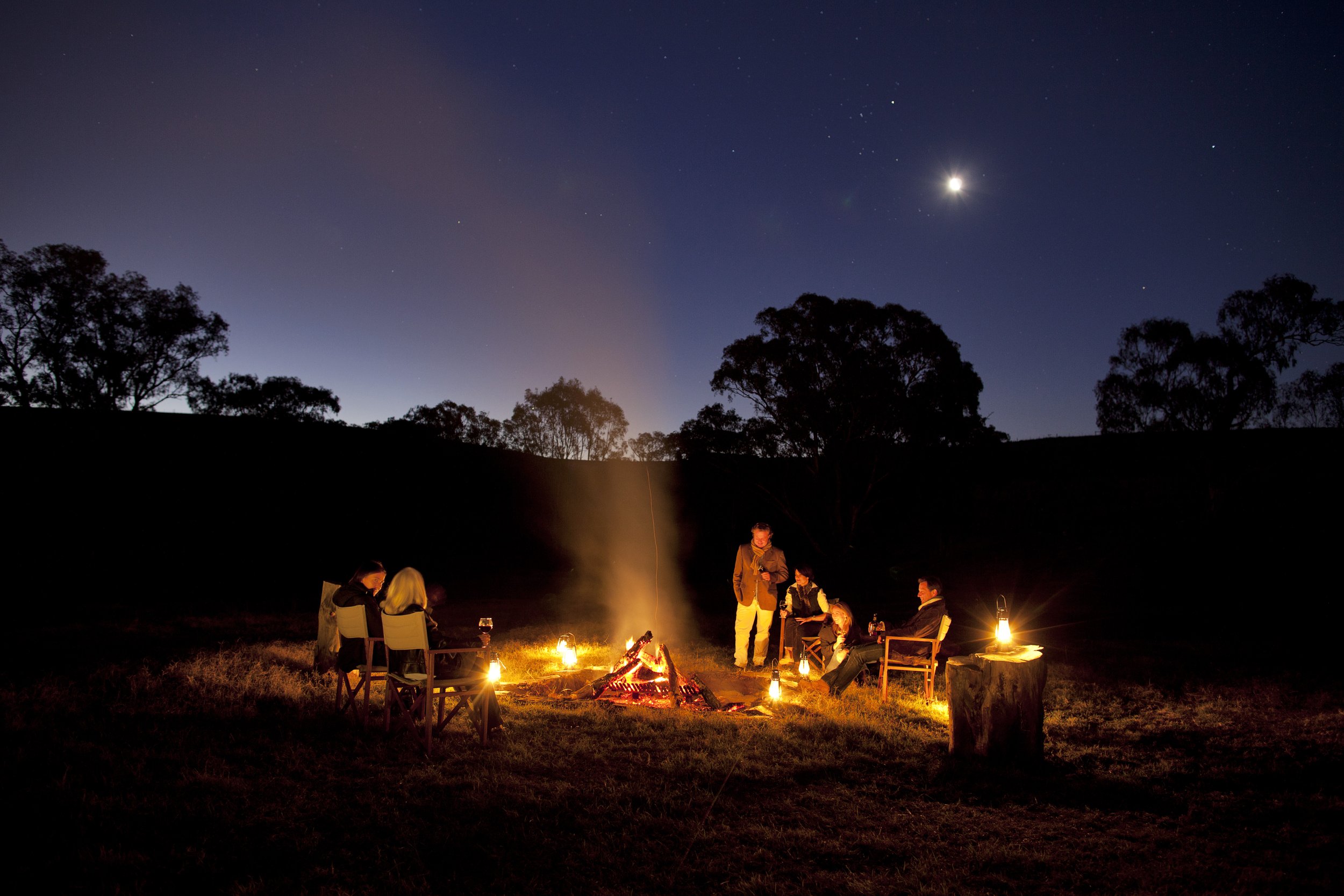 People gathered around a campfire under a starry night sky with the moon visible, in an outdoor setting with trees in the background.