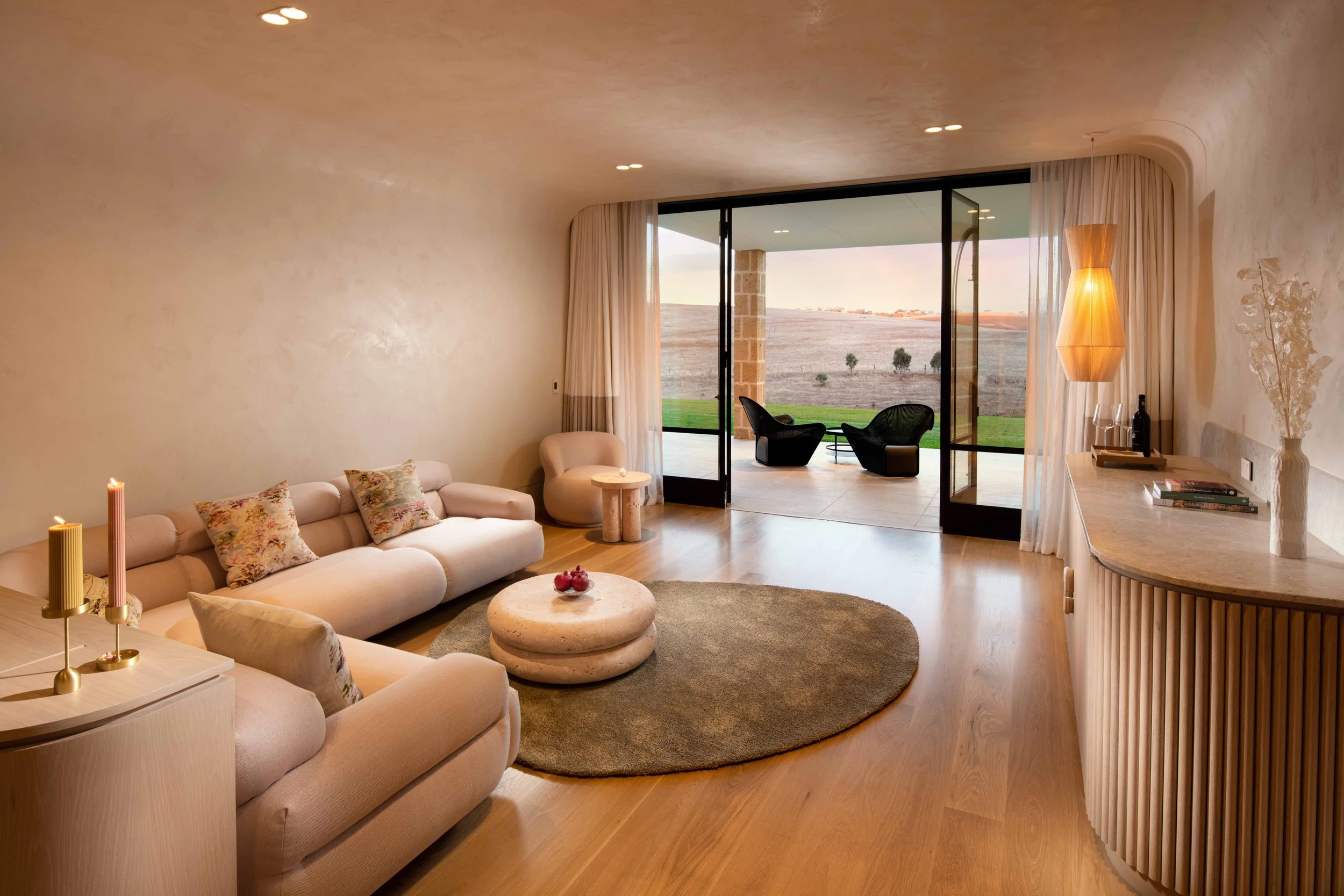 Living room with beige sofa and floral pillows, round stone coffee table with red apples, candles on side table, large sliding glass door opening to patio with two black chairs and view of fields and trees outside.
