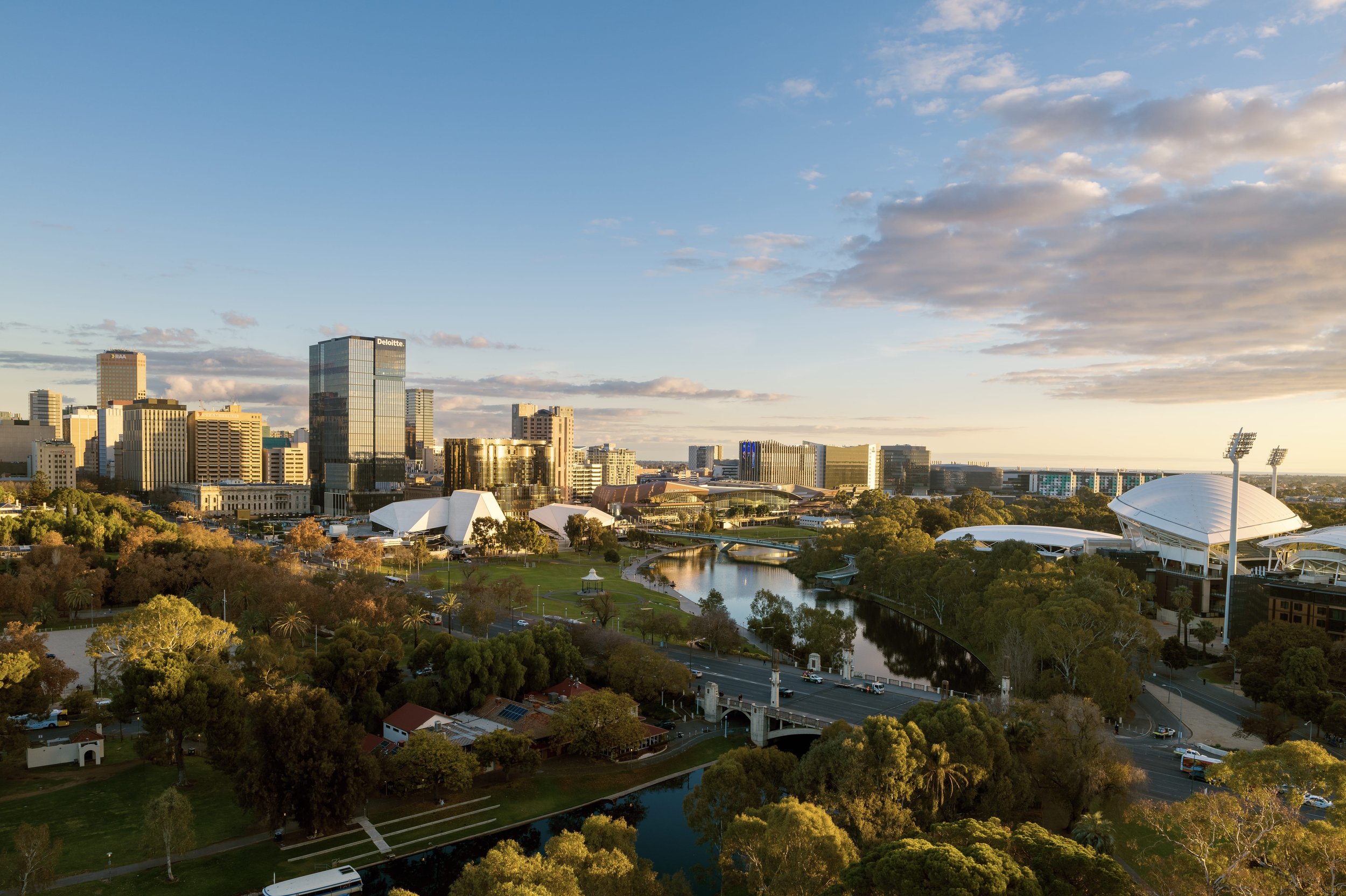 A city skyline during sunset with modern buildings, a river, a park, and a sports stadium.