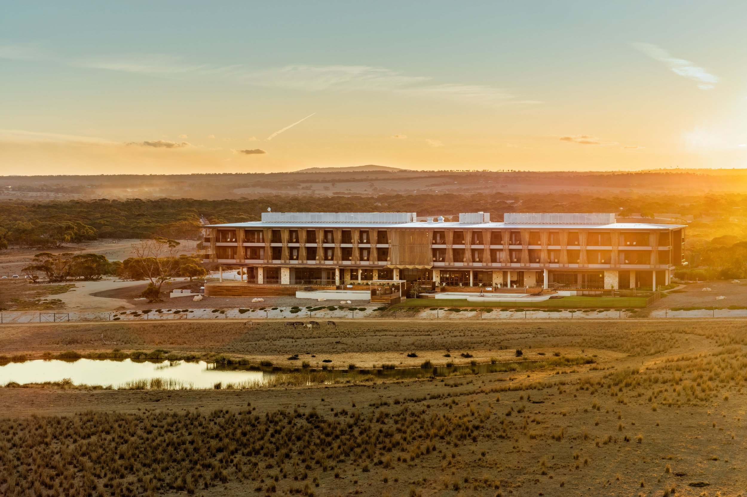 Modern building with large horizontal structure situated in a rural landscape during sunset, with hills and trees in the background and reflective water in the foreground.
