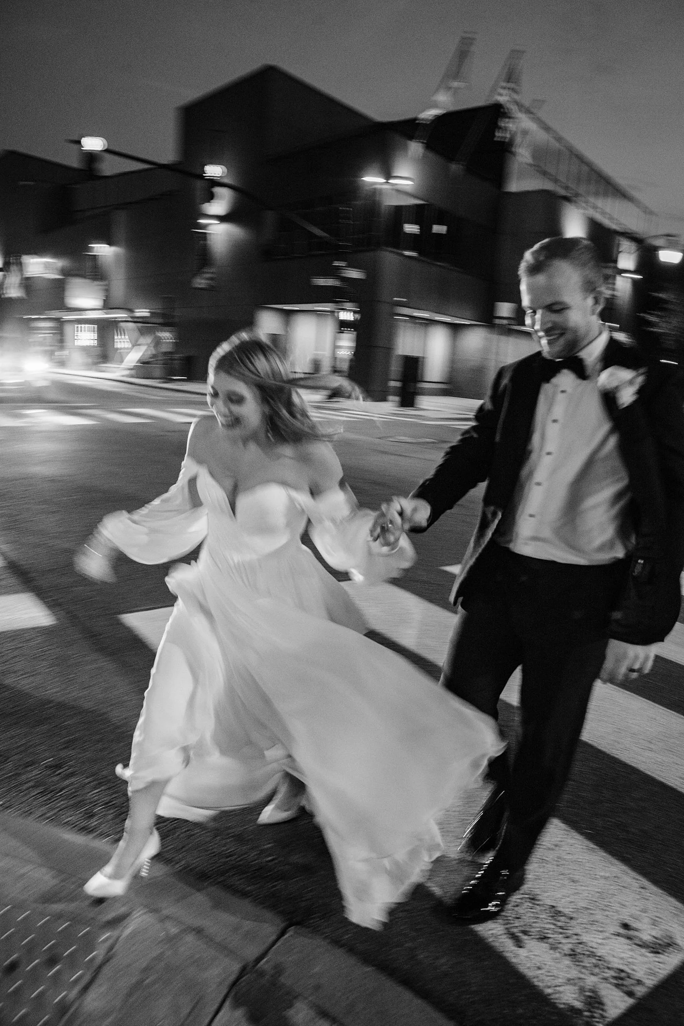 Black and white editorial style photo of a couple in wedding attire crossing a Providence, RI street at night. The woman is in a flowing wedding dress, and the man is in a tuxedo. They are smiling and holding hands, appearing joyful and playful.