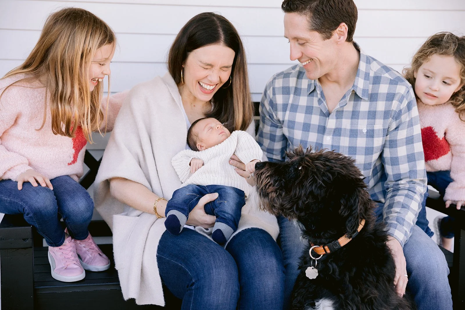 candid family photo of a family gathered on a bench outside, smiling and looking at a sleeping newborn held by an older woman, with two young girls and a dog nearby.
