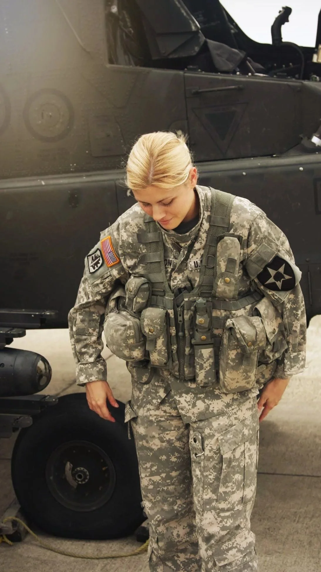 A female soldier in camouflage uniform stands in front of a Black Hawk helicopter, looking down with her head bowed and hands at her sides.