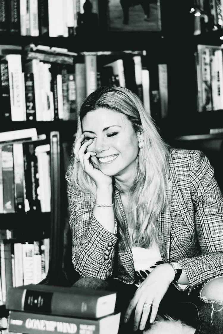 Black and white photo of a woman smiling and laughing in a bookstore, sitting in front of a bookshelf filled with books, wearing a plaid blazer and a watch.