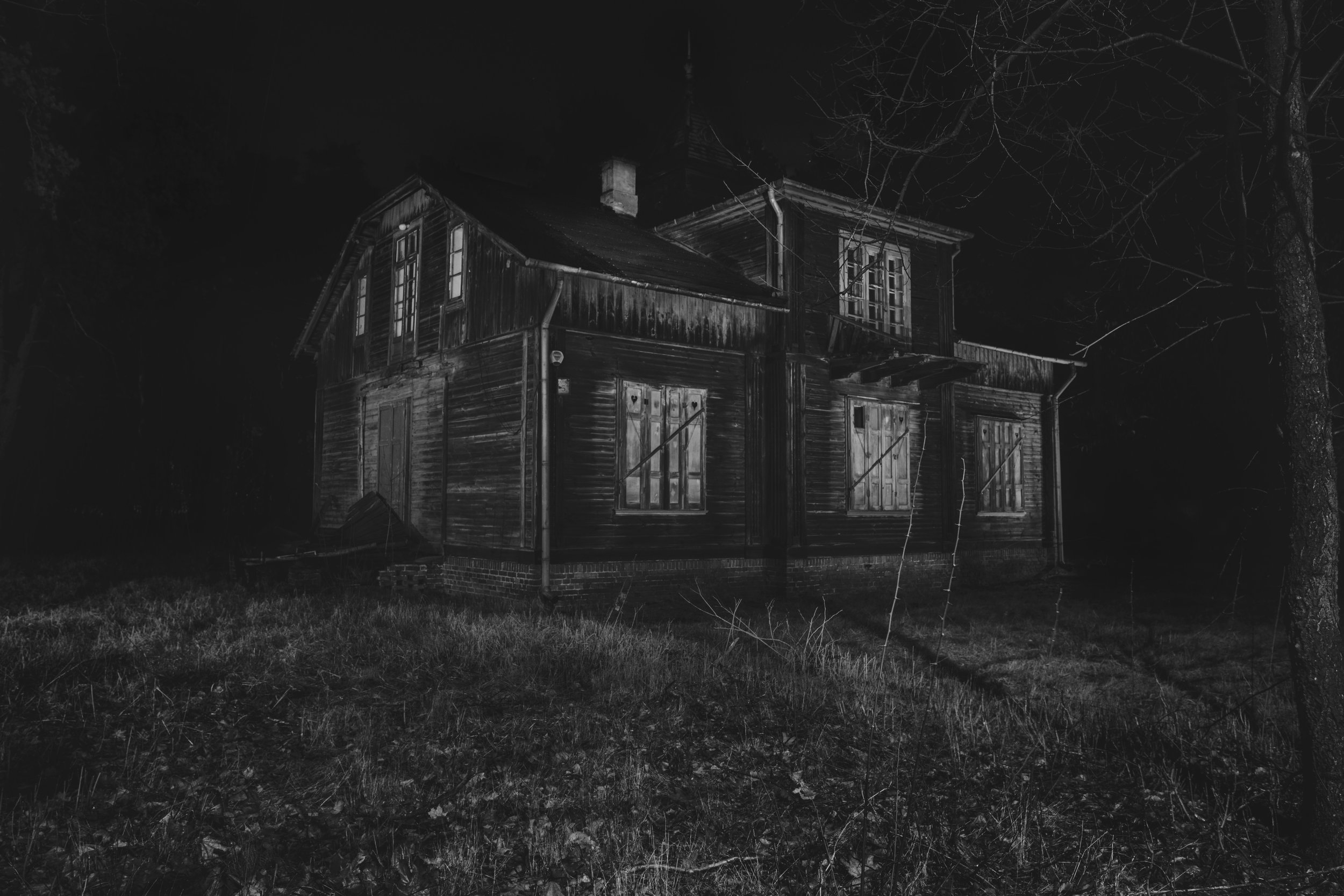 A dark, abandoned wooden house with boarded-up windows, situated in a grassy area with leafless trees surrounding it, under a night sky.