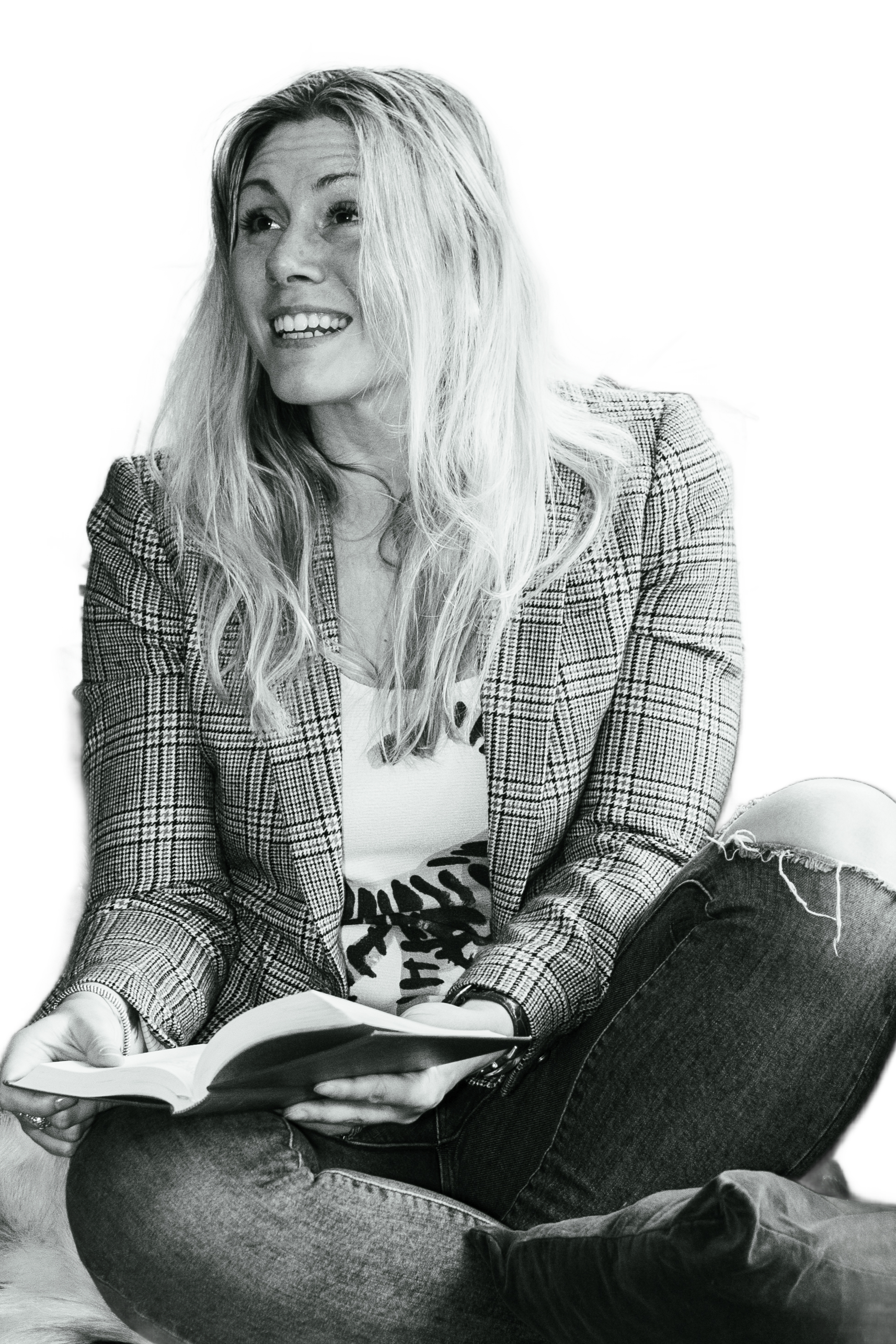 A woman with long blonde hair smiling while sitting cross-legged and holding an open book, wearing a plaid blazer and ripped jeans.