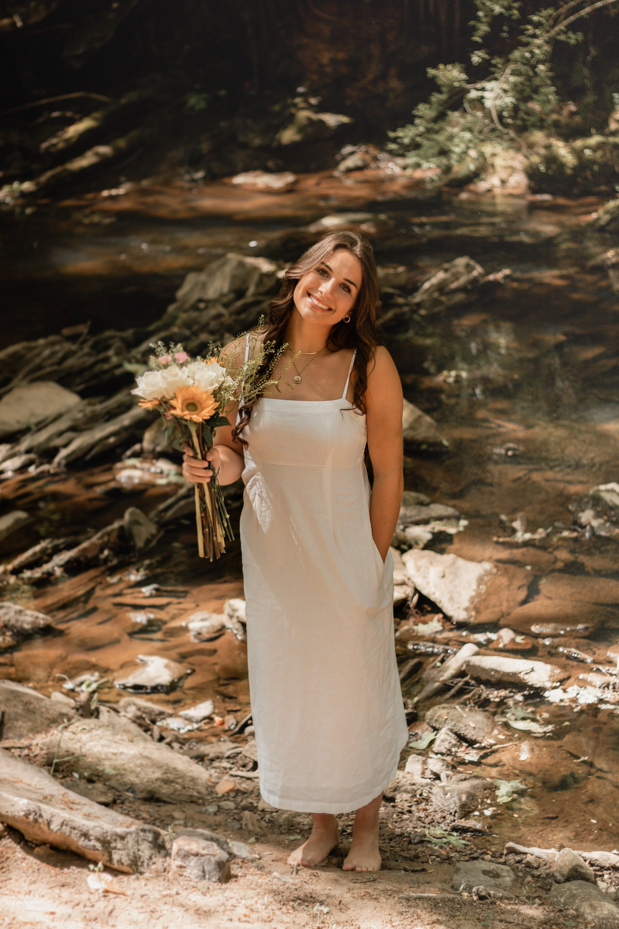 Senior portraits by a creek/river. Girl in a long white dress with flowers smiling by a creek/river.