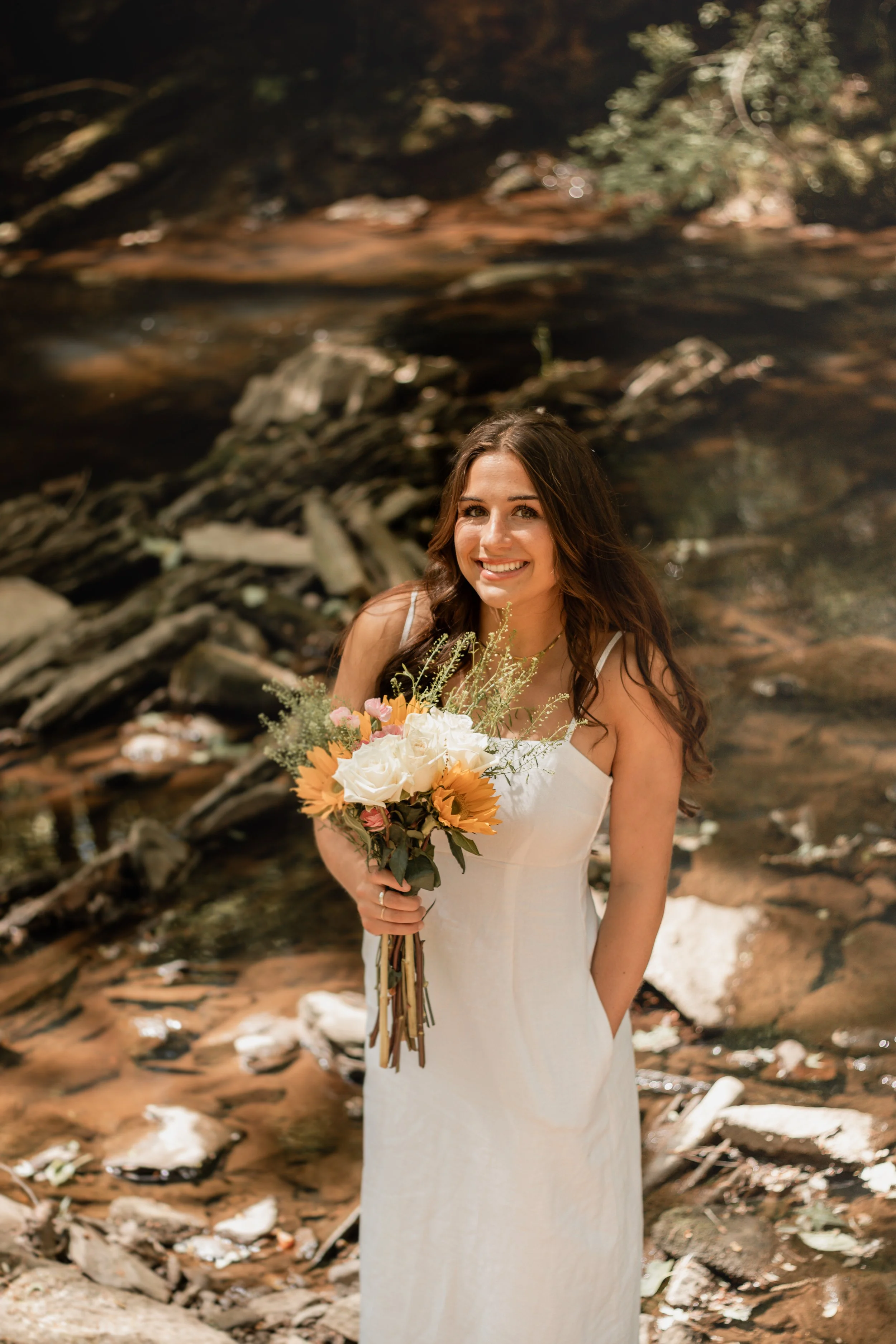 Senior portraits by a creek/river. Girl in a long white dress with flowers smiling by a creek/river.