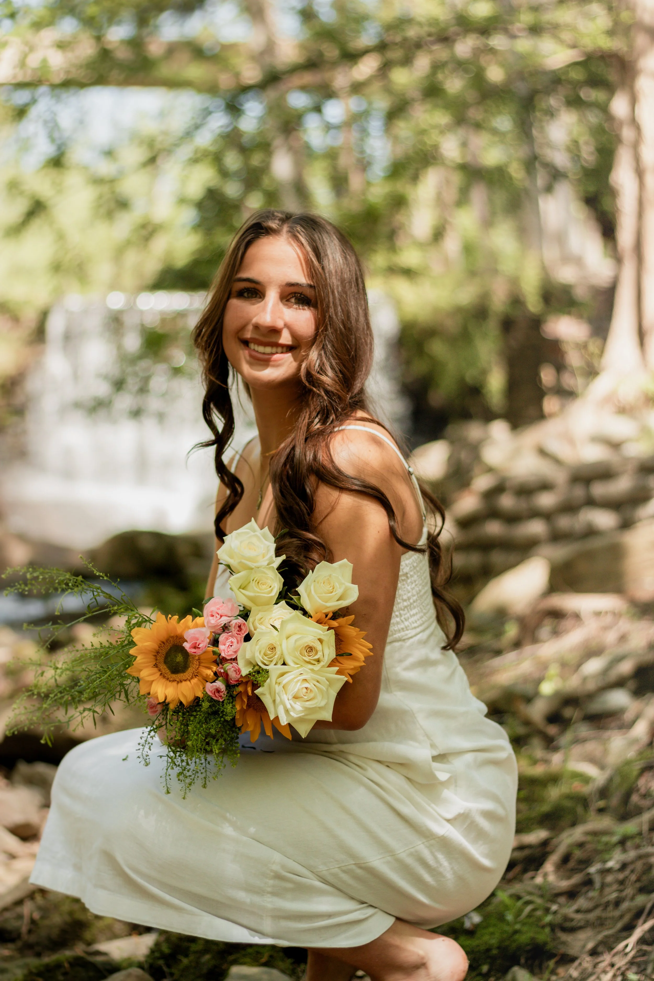 Senior portraits by a creek/river. Girl in a long white dress with flowers smiling by a creek/river.