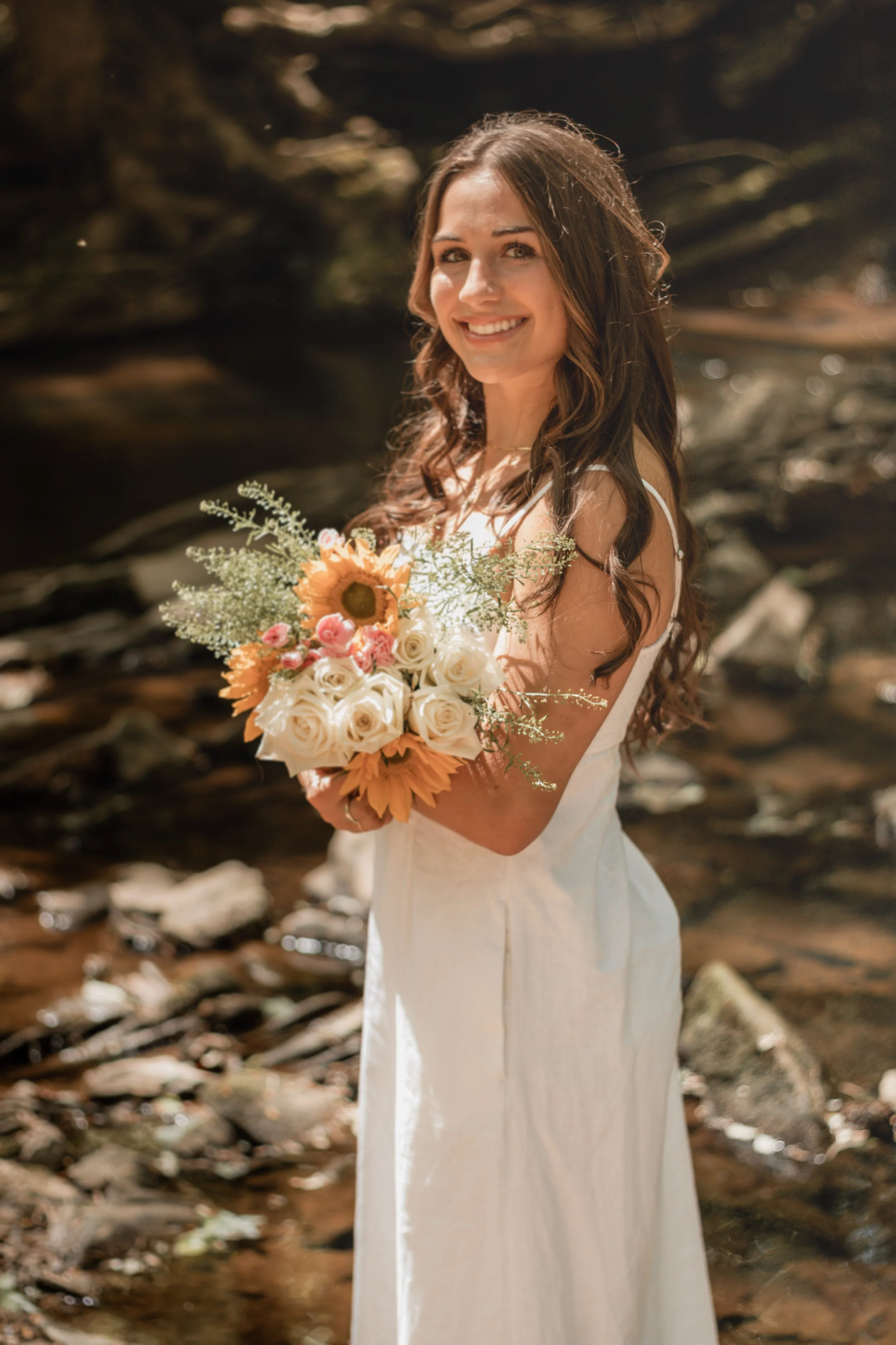 Senior portraits by a creek/river. Girl in a long white dress with flowers smiling by a creek/river.