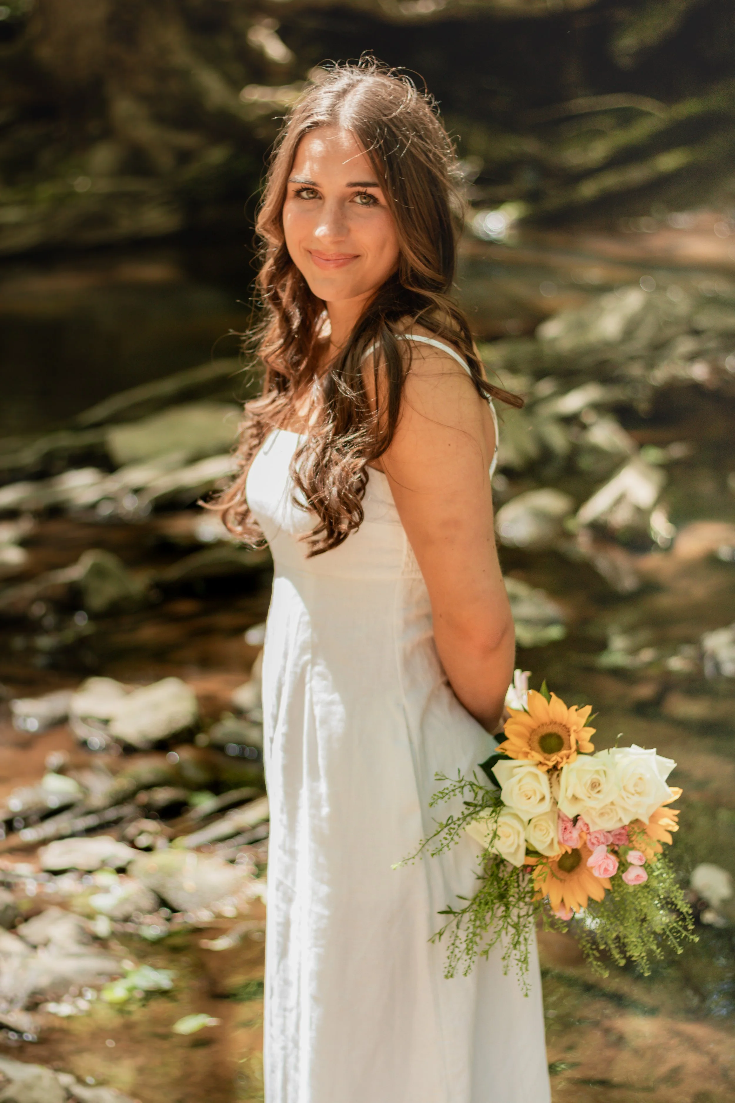 Senior portraits by a creek/river. Girl in a long white dress with flowers smiling by a creek/river.