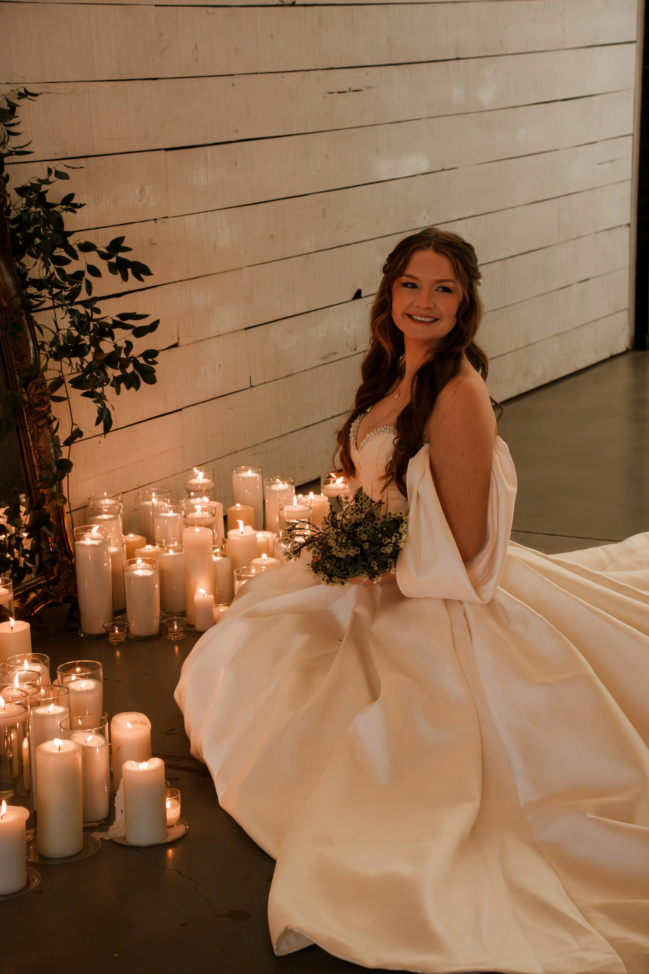 Bride sitting in front of mirror and candles in her wedding dress with her bouquet in her arms