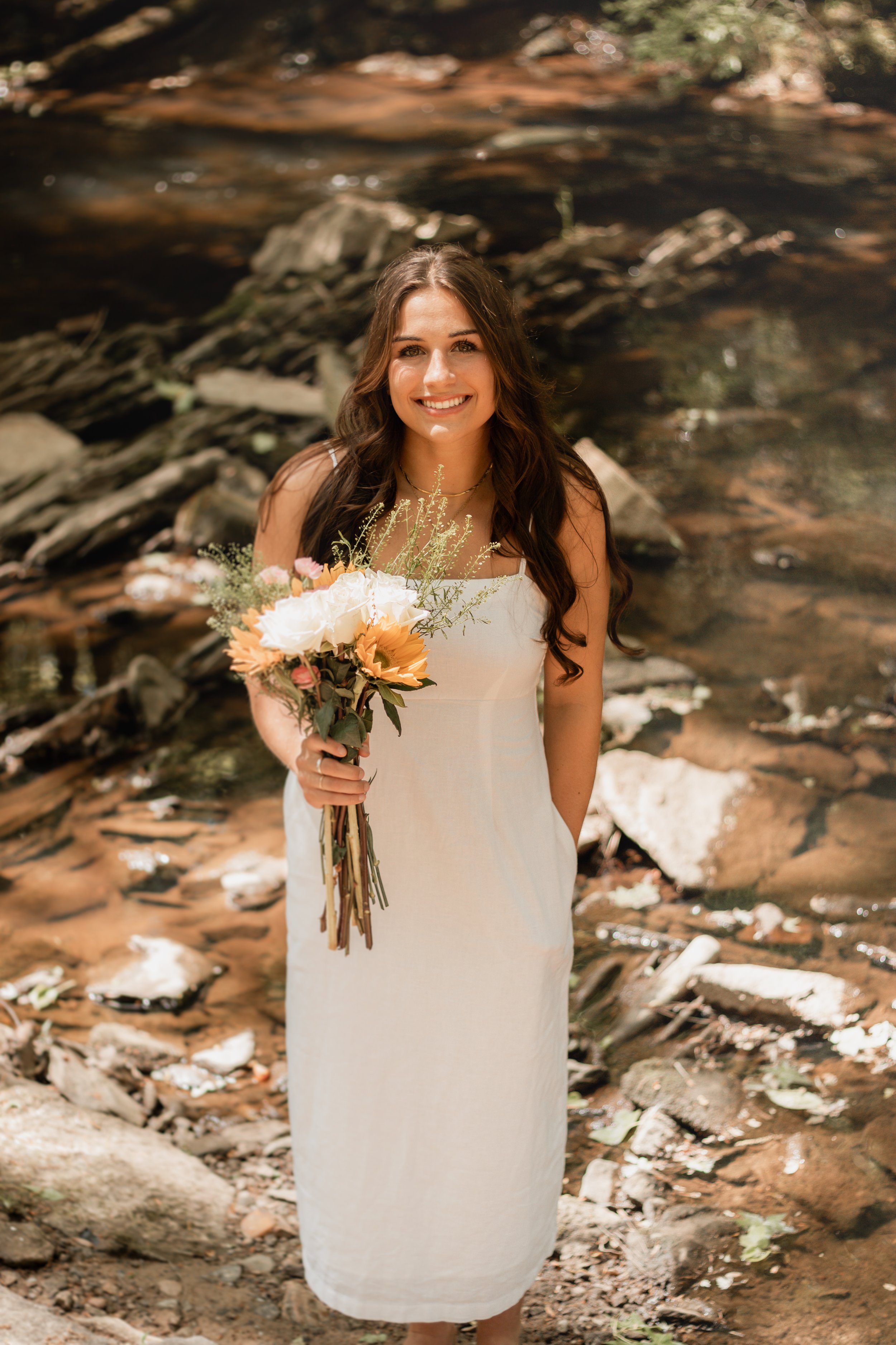 Senior portraits by a creek/river. Girl in a long white dress with flowers smiling by a creek/river.