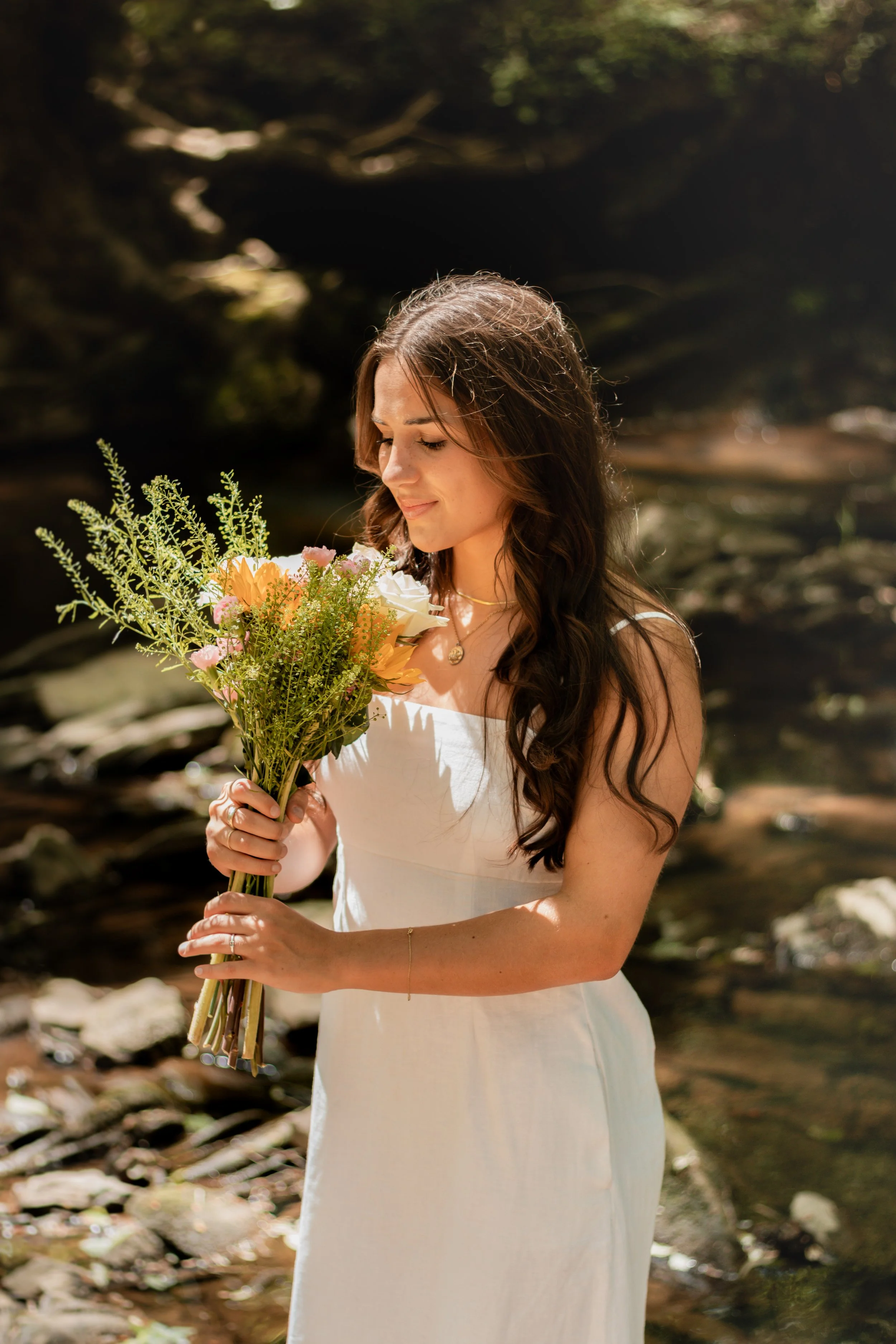 Senior portraits by a creek/river. Girl in a long white dress with flowers smiling by a creek/river.