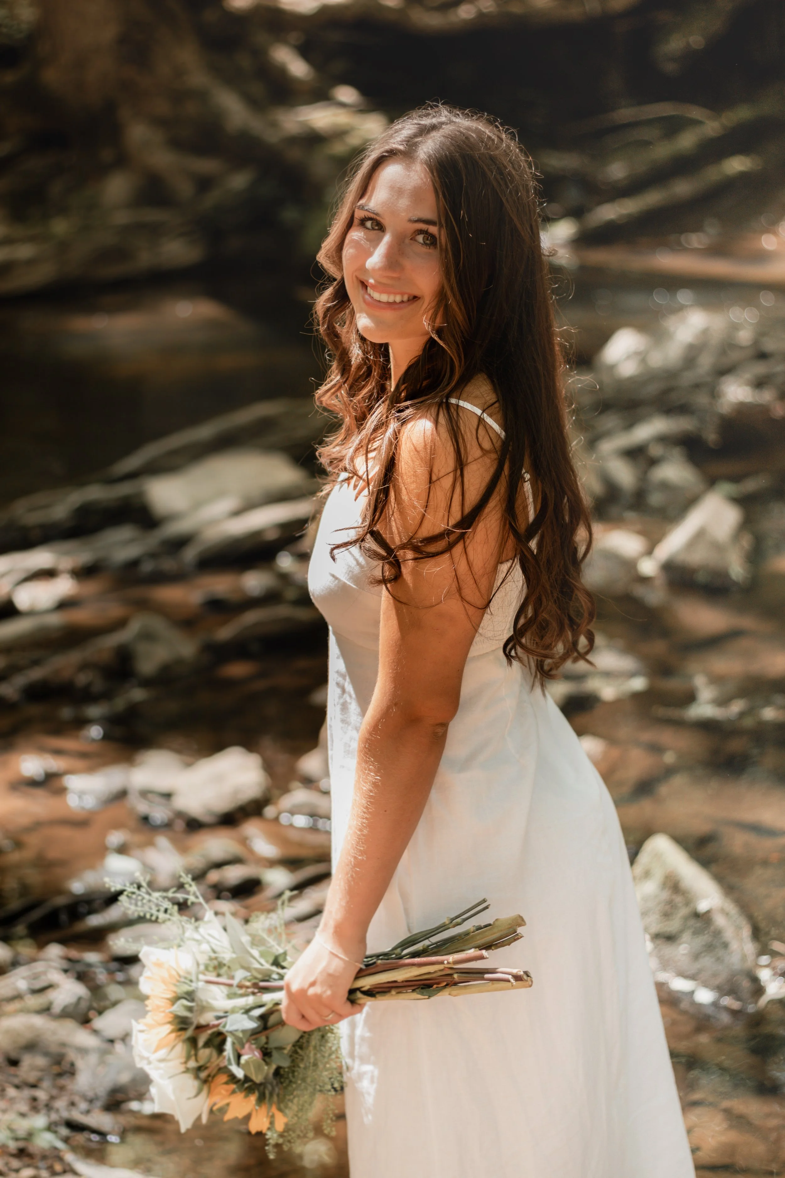 Senior portraits by a creek/river. Girl in a long white dress with flowers smiling by a creek/river.
